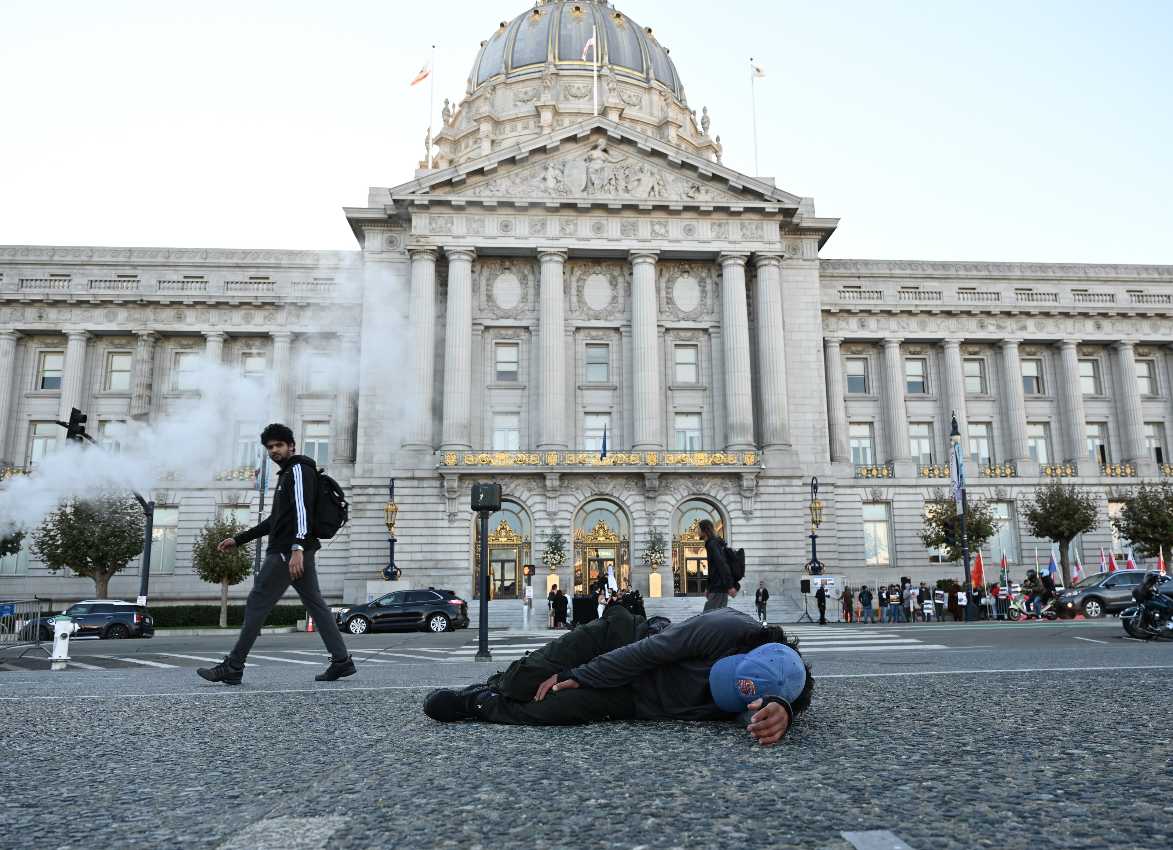 A person lies on the street in front of a large, ornate building, while another person walks past. Steam or smoke is billowing nearby.