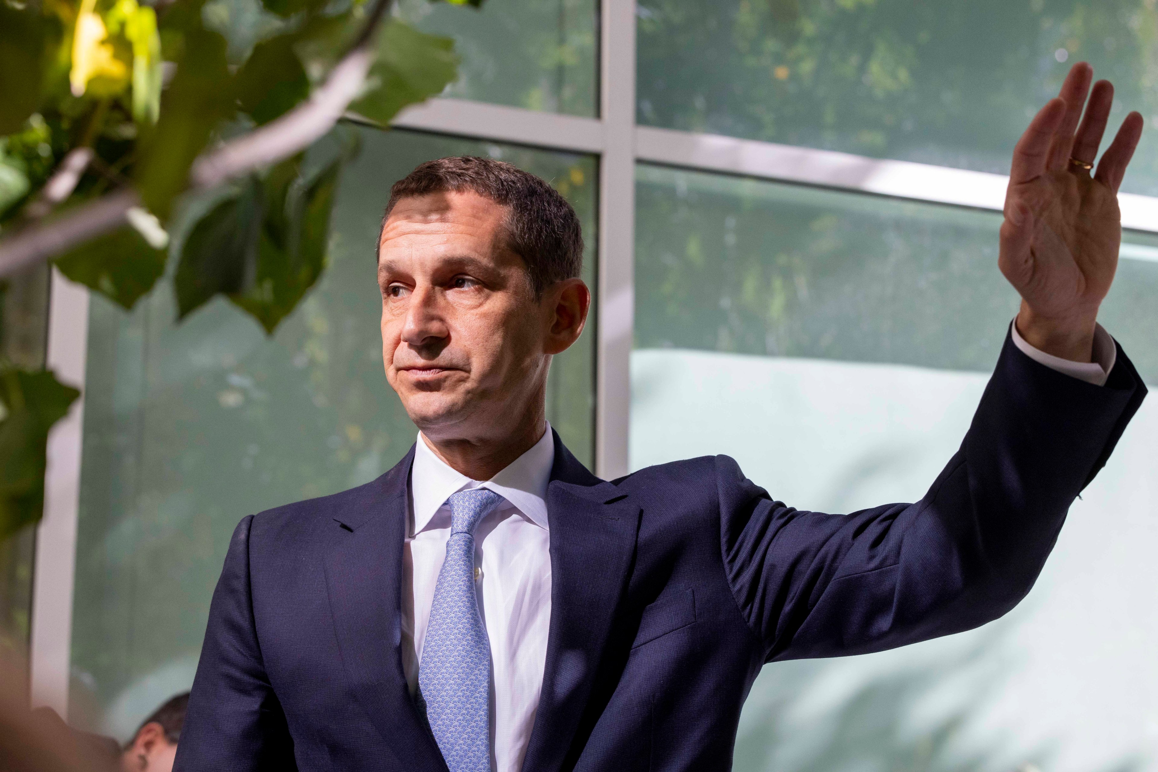 A man in a suit stands indoors, his right arm raised. Green leaves partially obscure the view, and large windows are in the background.