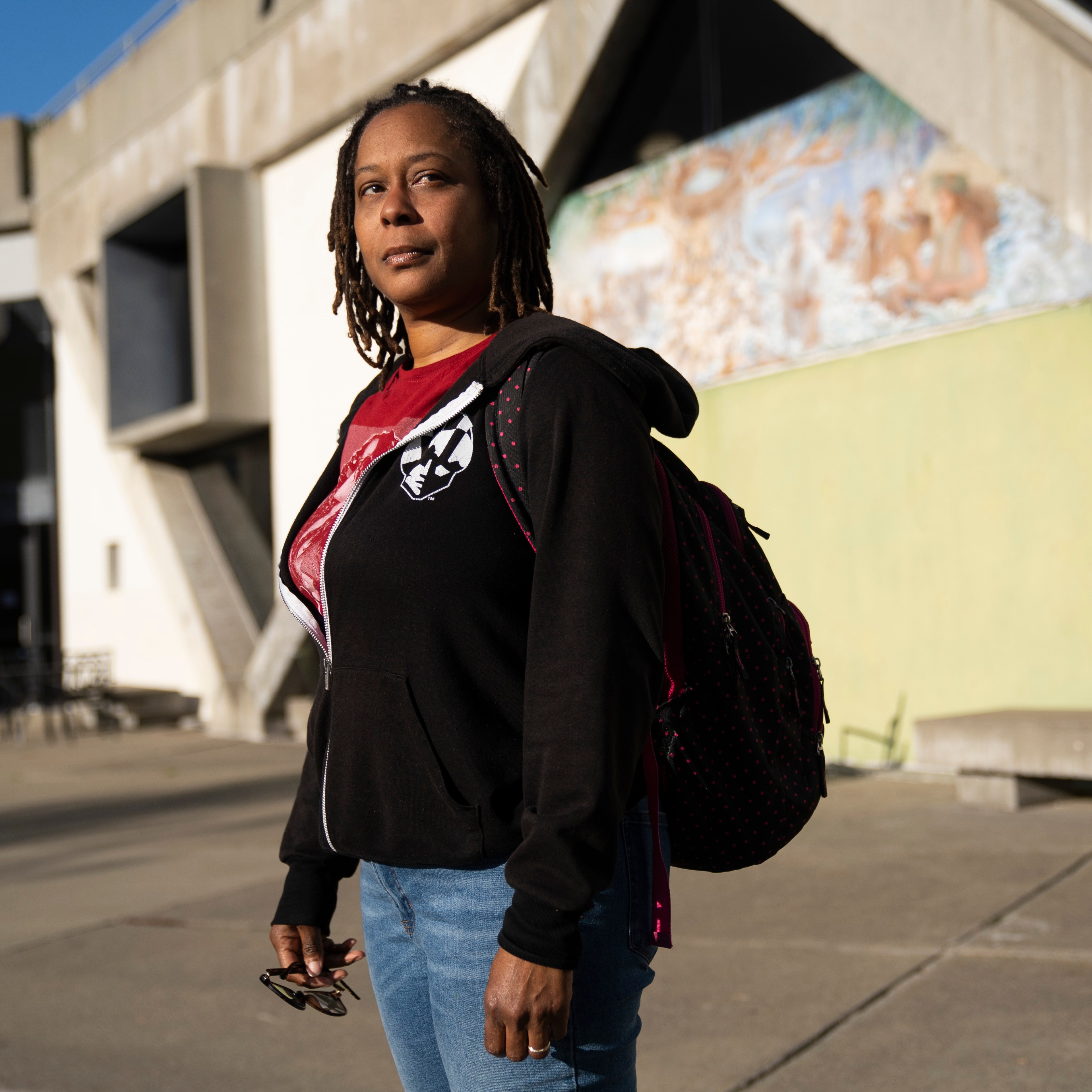 A person with dreadlocks, wearing a black hoodie and blue jeans, stands outside a building with geometric concrete architecture, under sunlight.