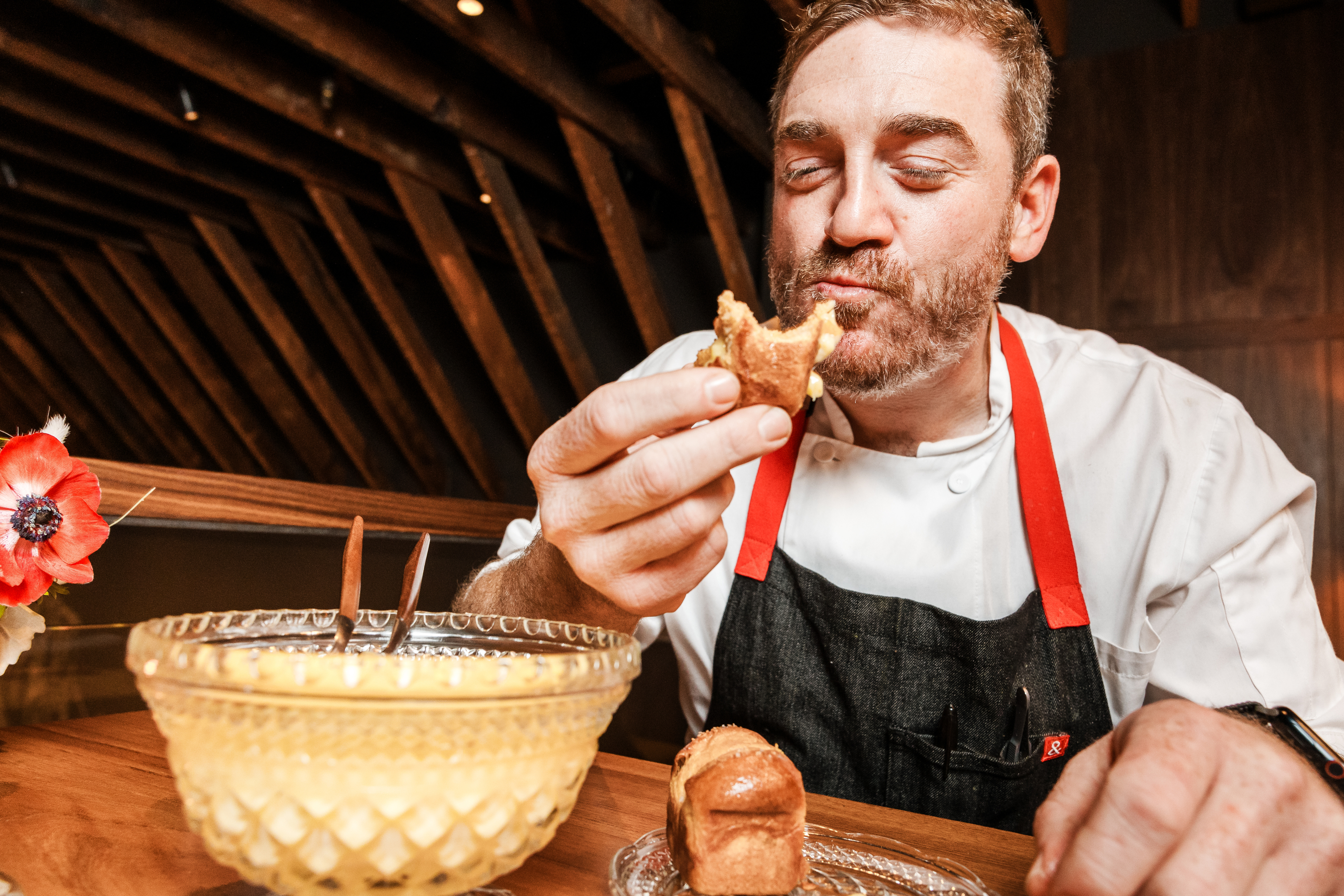 A chef in a white shirt and apron enjoys a bite of pastry. He sits at a wooden table with a decorative bowl, a small loaf, and a red flower in view.