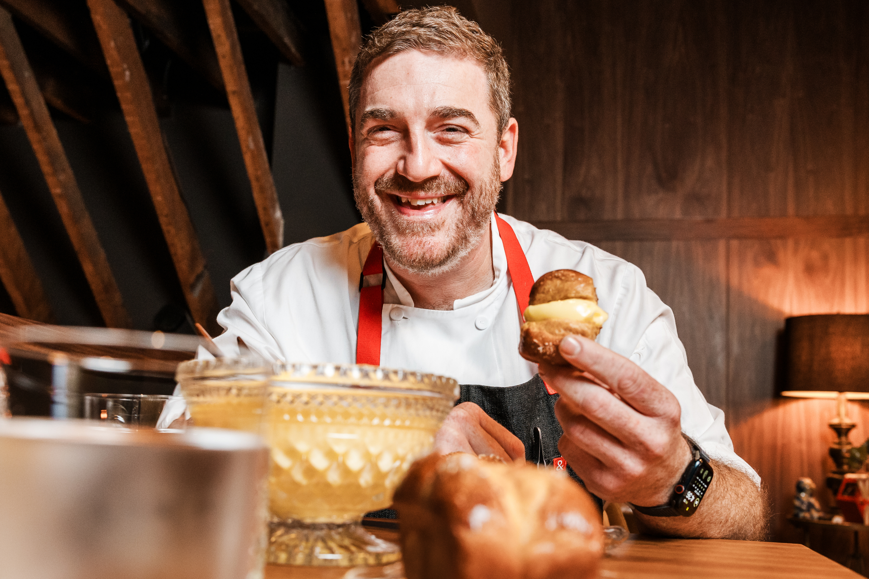 A smiling chef in a white shirt and red apron holds a pastry filled with cream, sitting at a table with a decorative bowl and a lamp in the background.