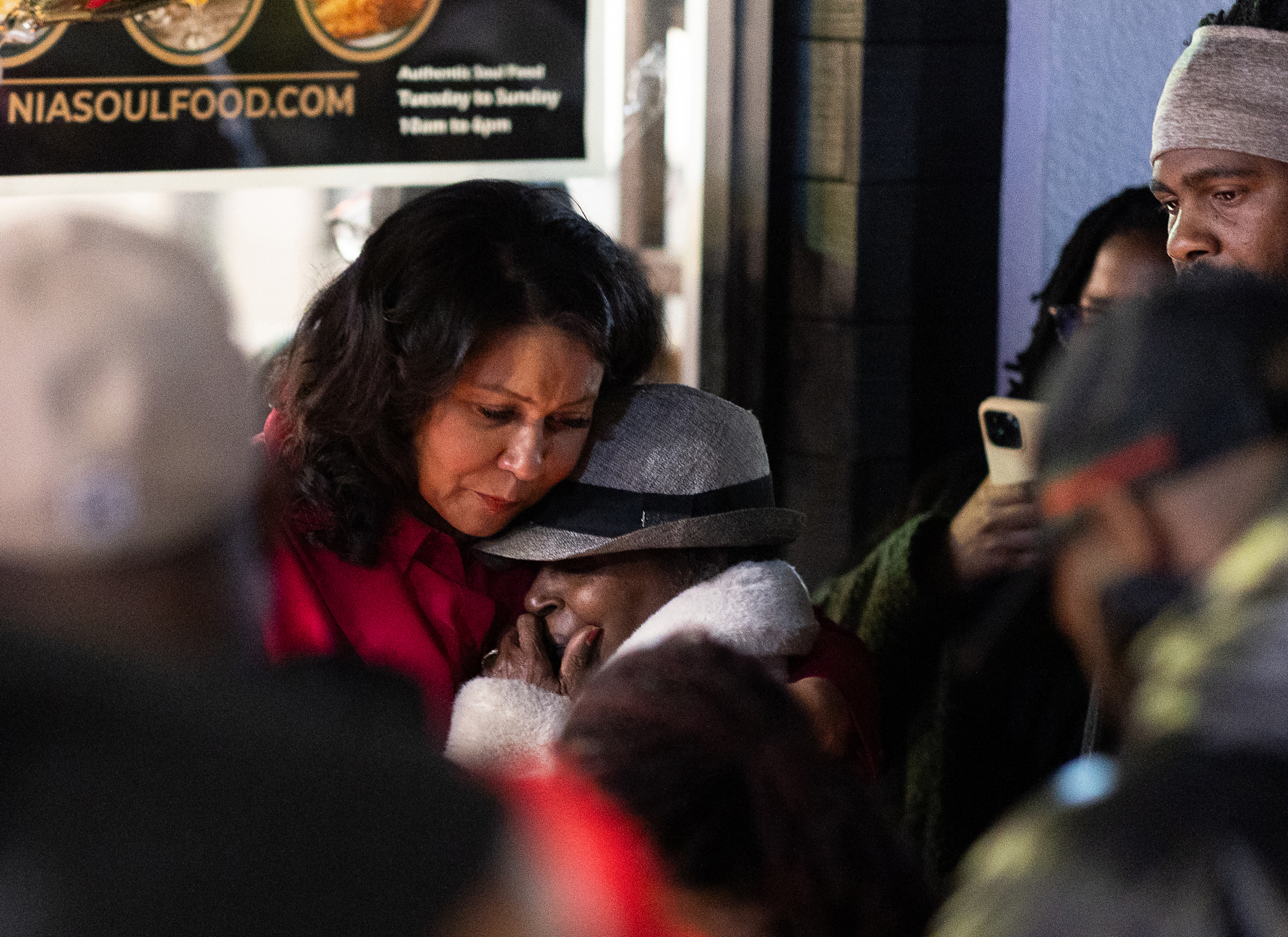 A woman in a red coat is comforting another, who is wearing a gray hat, amidst a group. They're standing outside a building, with an emotional atmosphere.