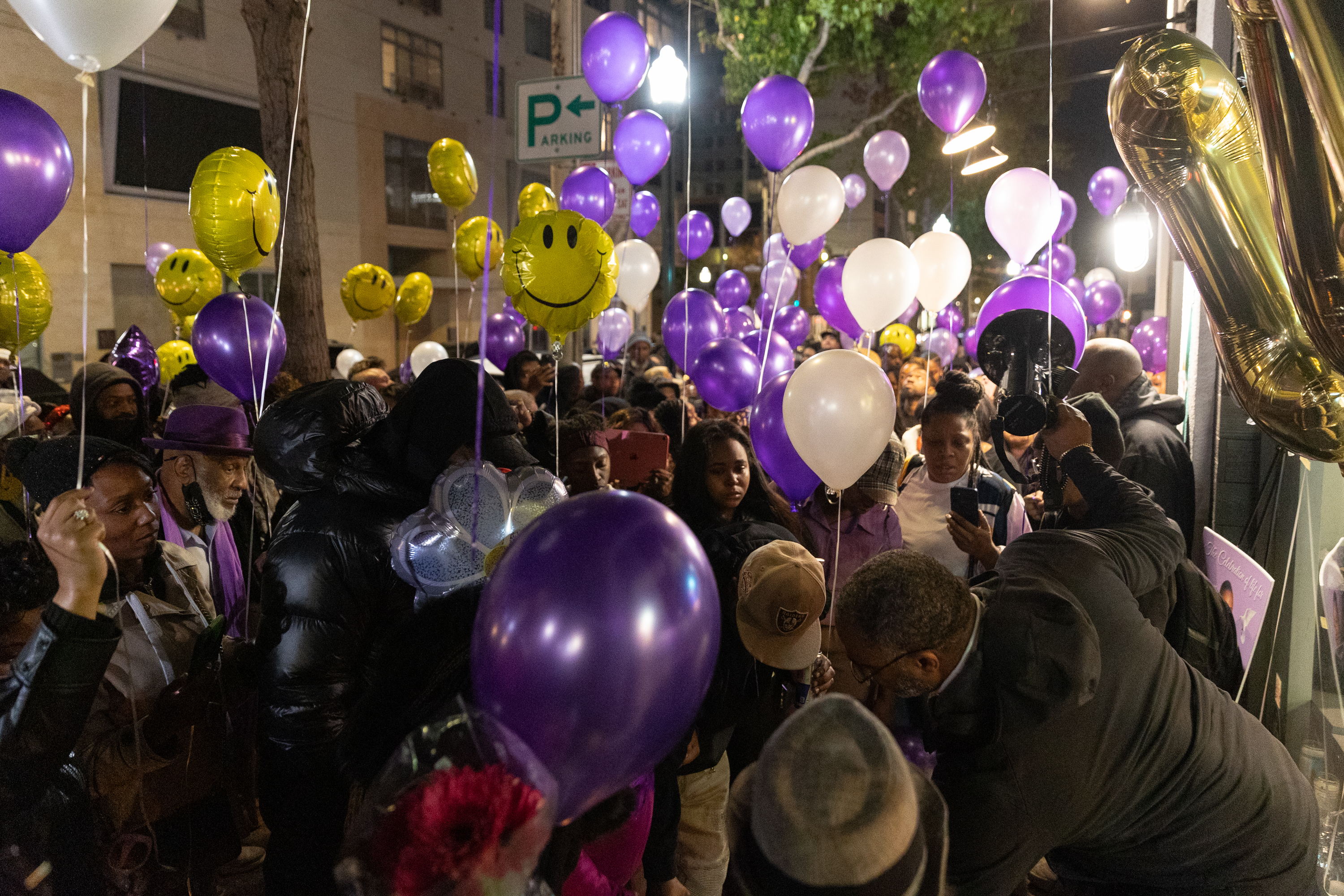 A crowd gathers at night holding purple, white, and smiley face balloons. They stand closely together, many capturing the moment on their phones.