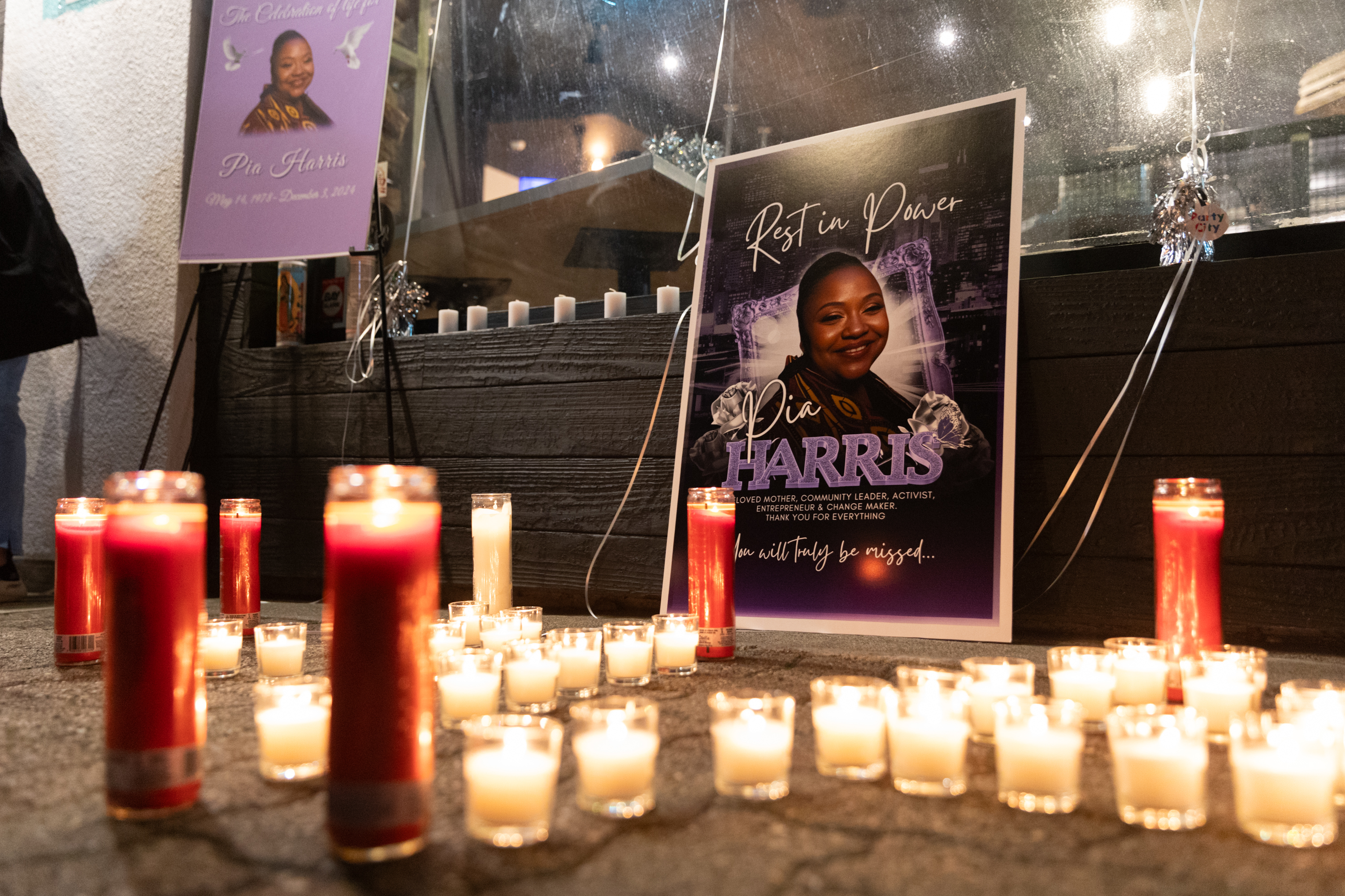 A memorial with lit candles surrounds posters honoring Pia Harris. The posters display her image, name, dates, and heartfelt tributes.