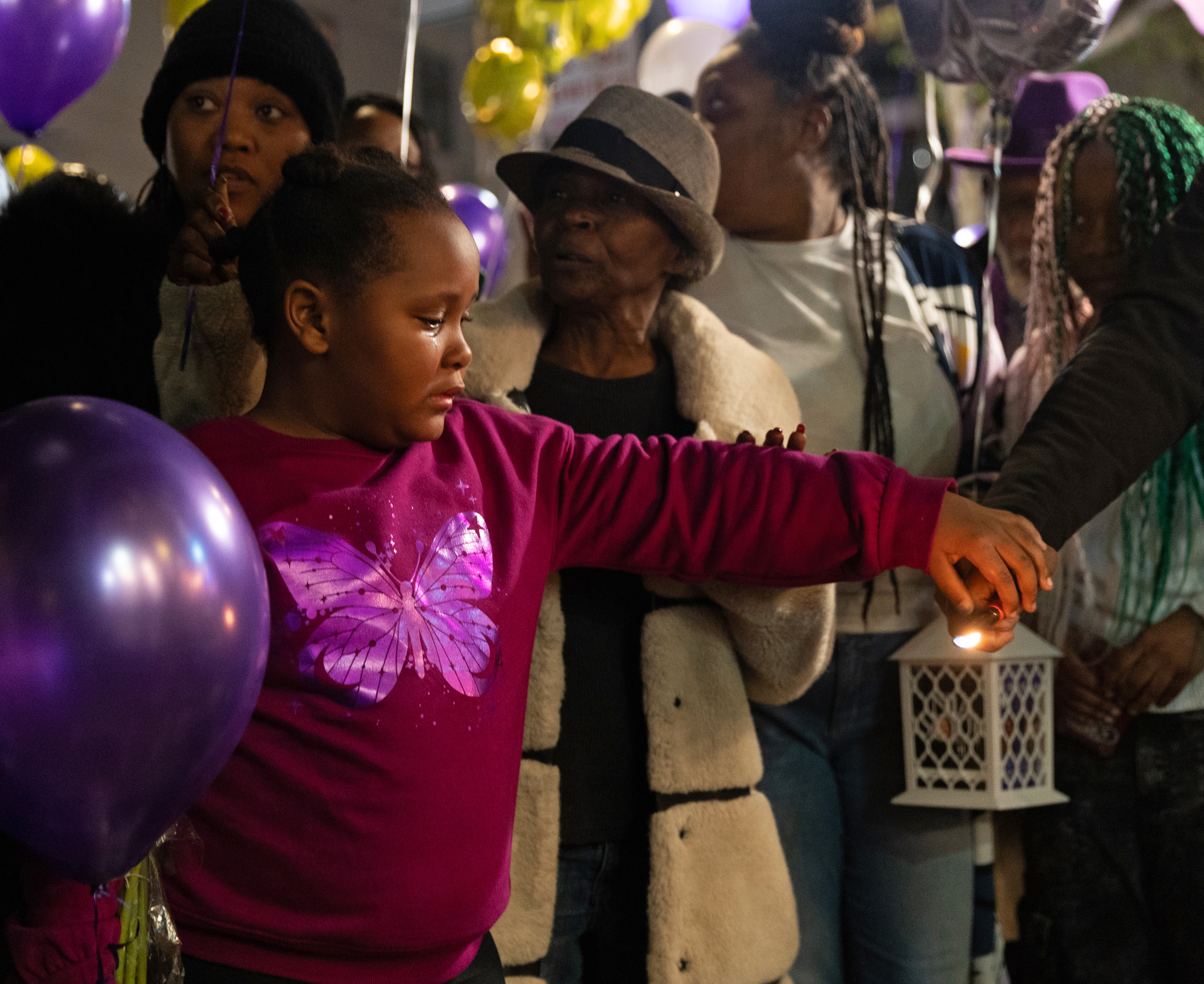 A young girl in a pink butterfly shirt holds a candle at a vigil, surrounded by people holding purple balloons. An older woman and others support her.