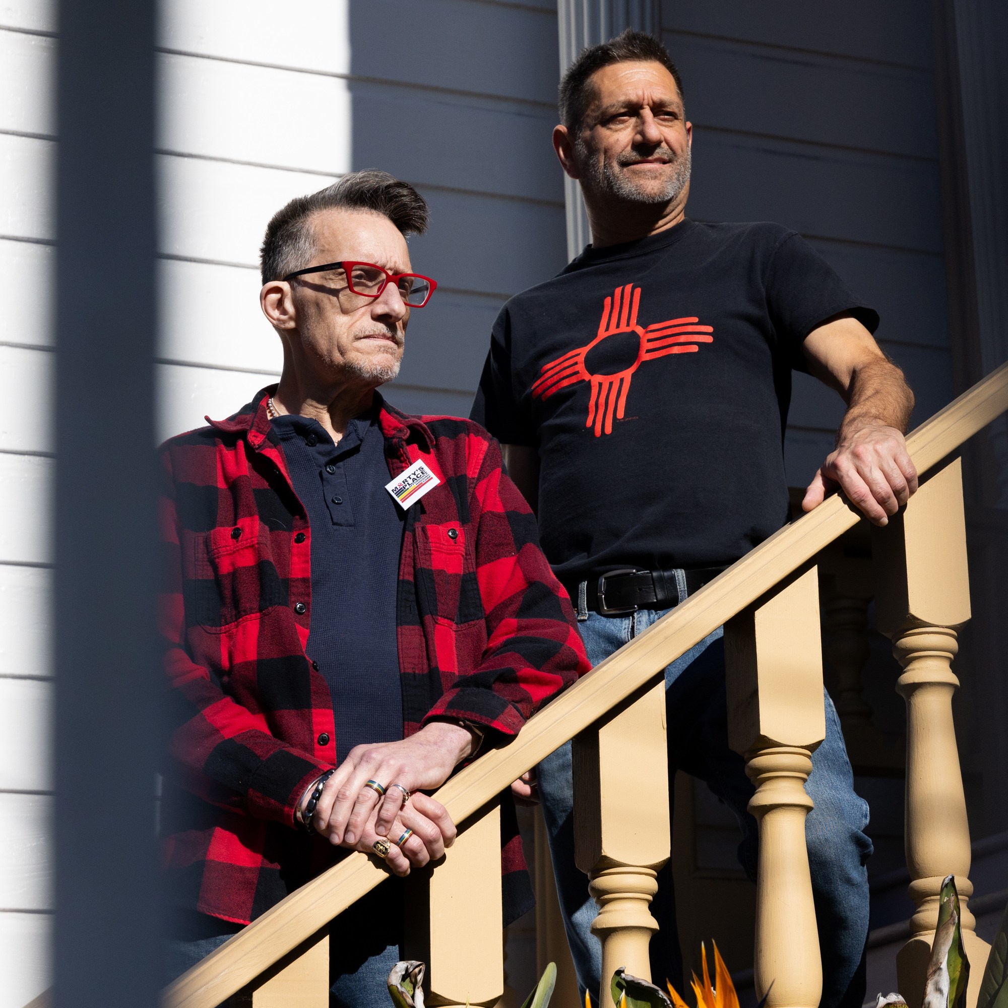 Two people stand on a wooden staircase. One wears a black and red checkered shirt and glasses, the other a black T-shirt with a red symbol. Sunlight casts shadows.