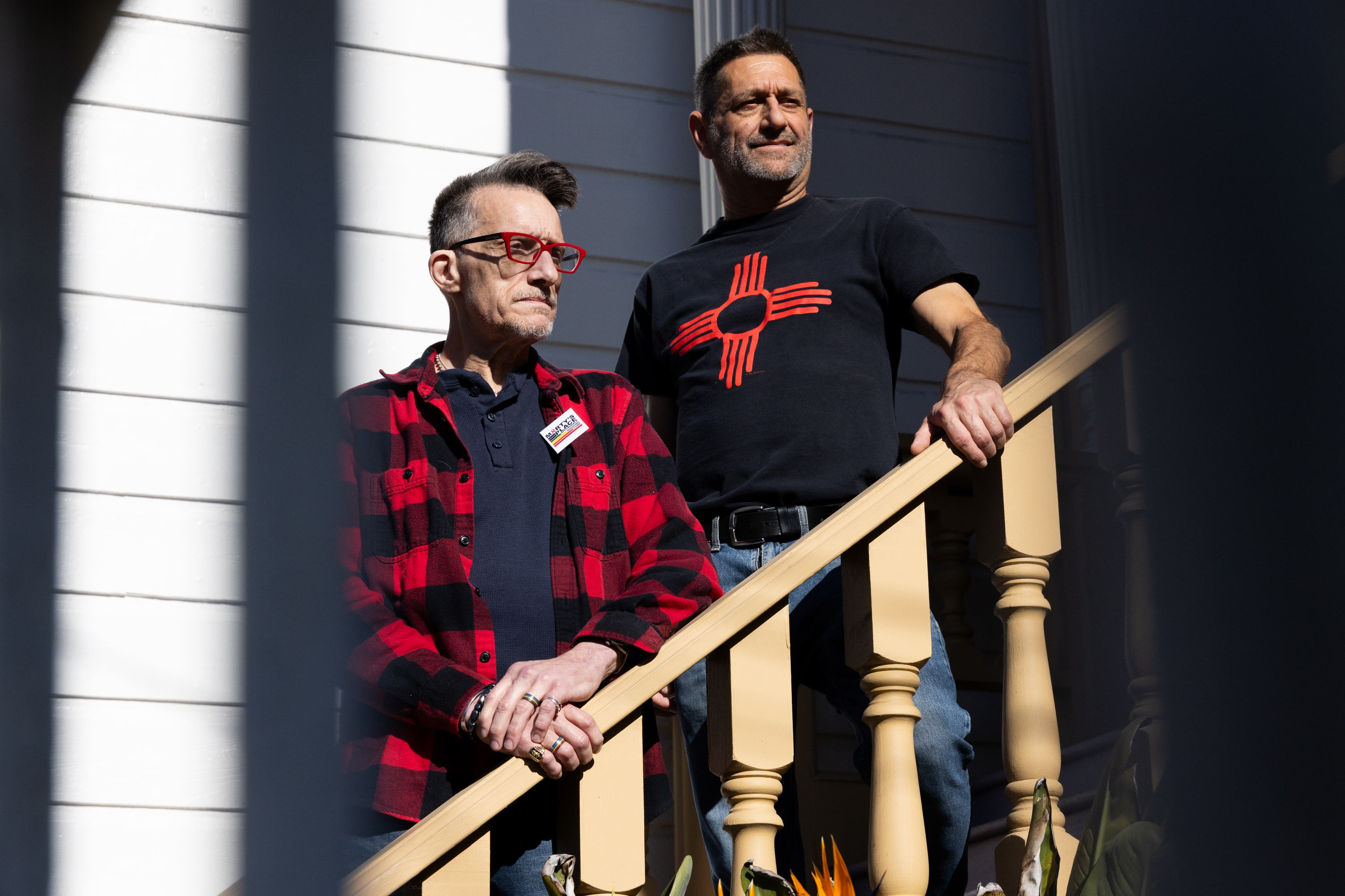 Two people stand on a wooden staircase. One wears a black and red checkered shirt and glasses, the other a black T-shirt with a red symbol. Sunlight casts shadows.