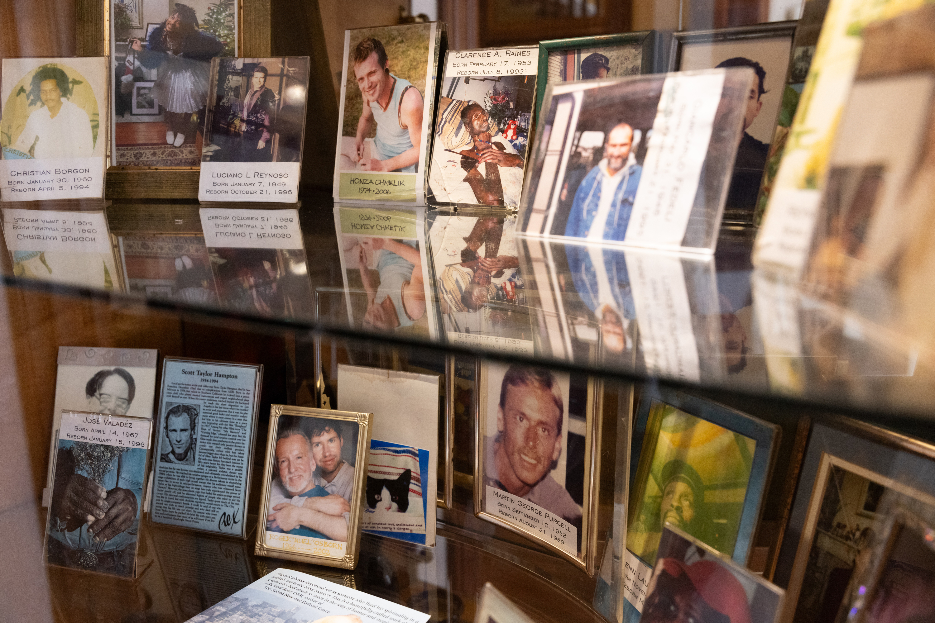 The image shows a glass display case filled with framed photographs and cards, each with names, birth and death dates, reflecting memorial tributes.