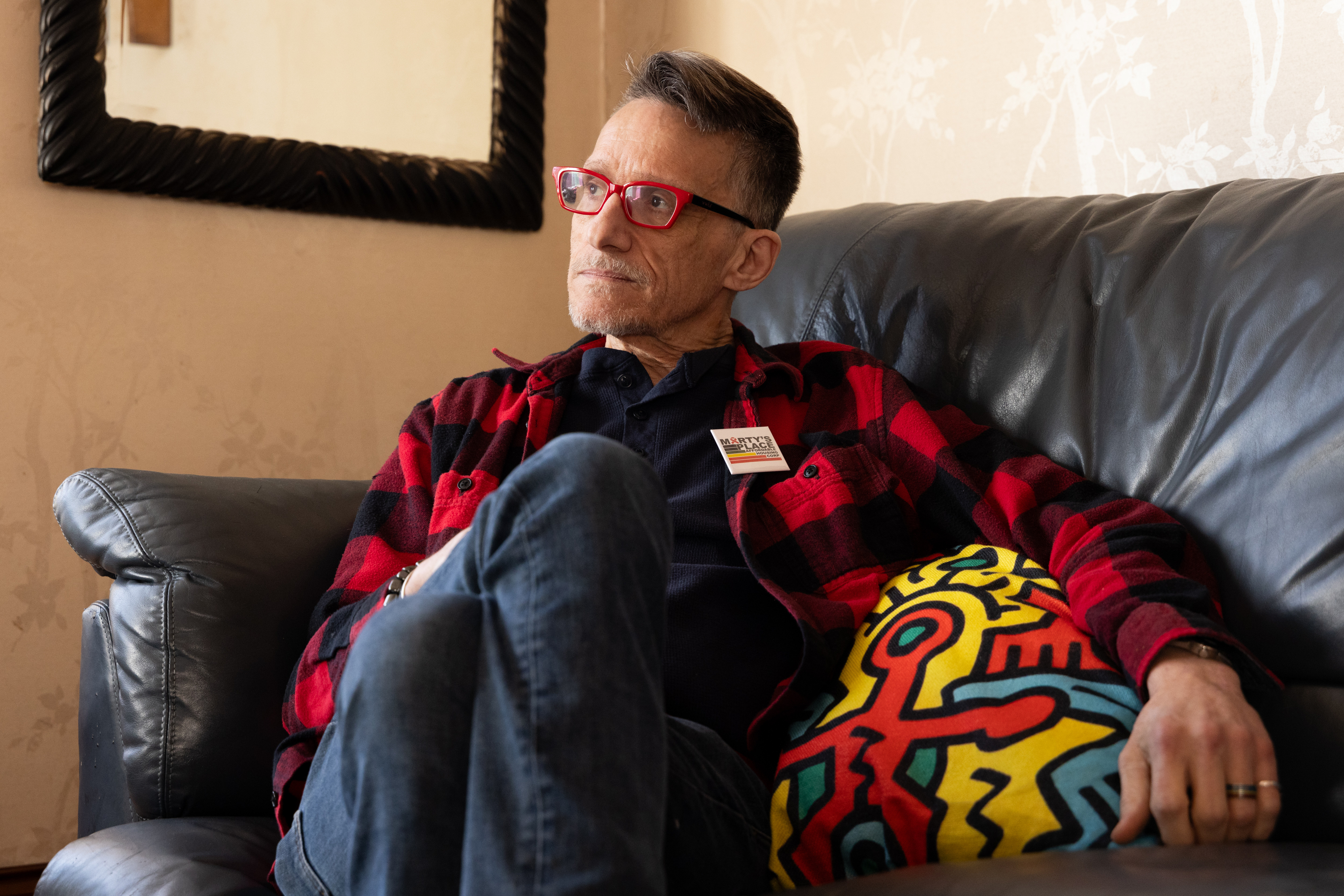 A person with red glasses and a red plaid shirt sits on a couch, holding a colorful pillow with an abstract design. A mirror is on the wall behind them.