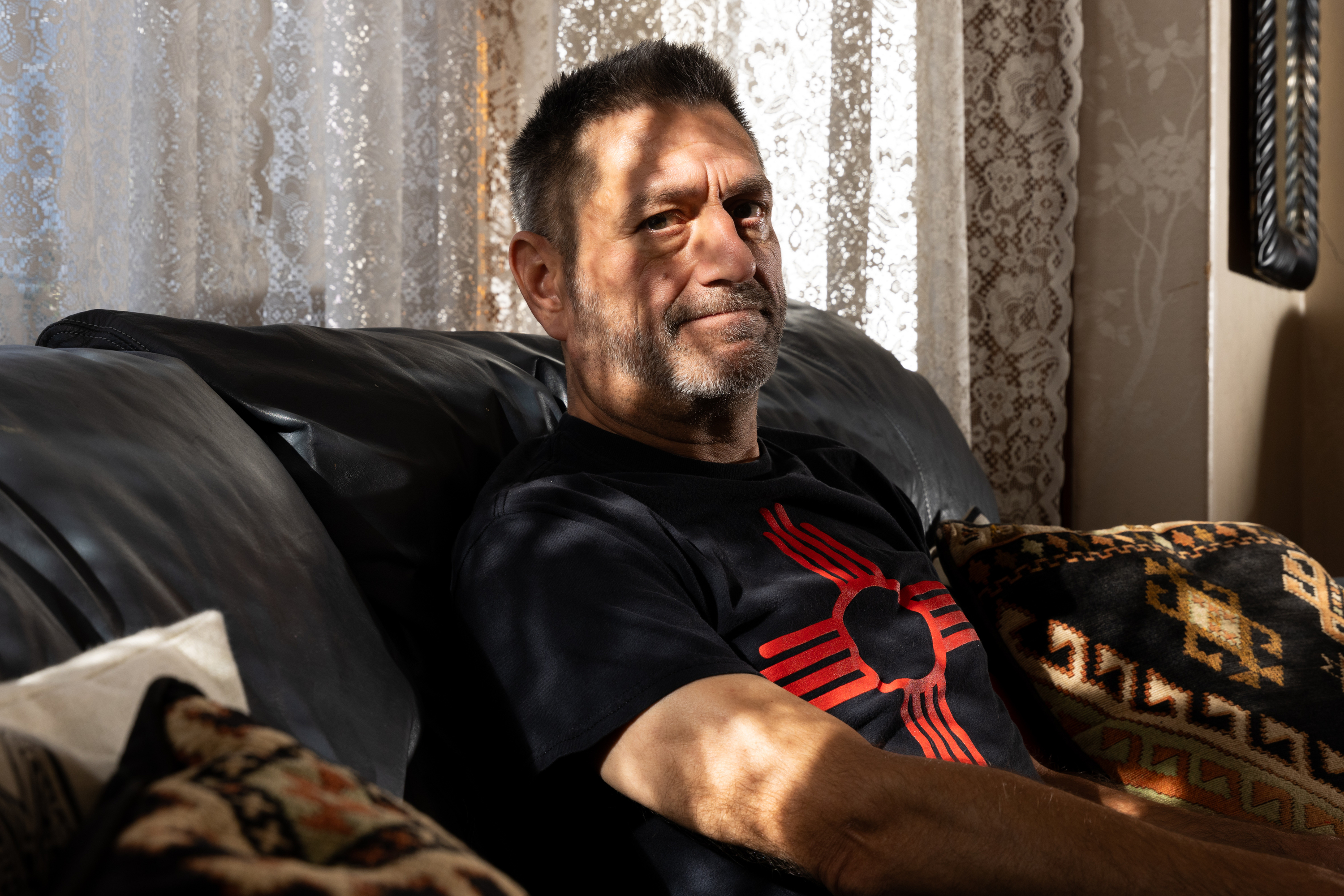 A man with short hair and a beard sits on a couch in a sunlit room, wearing a black shirt with a red symbol. Lace curtains and patterned pillows are visible.