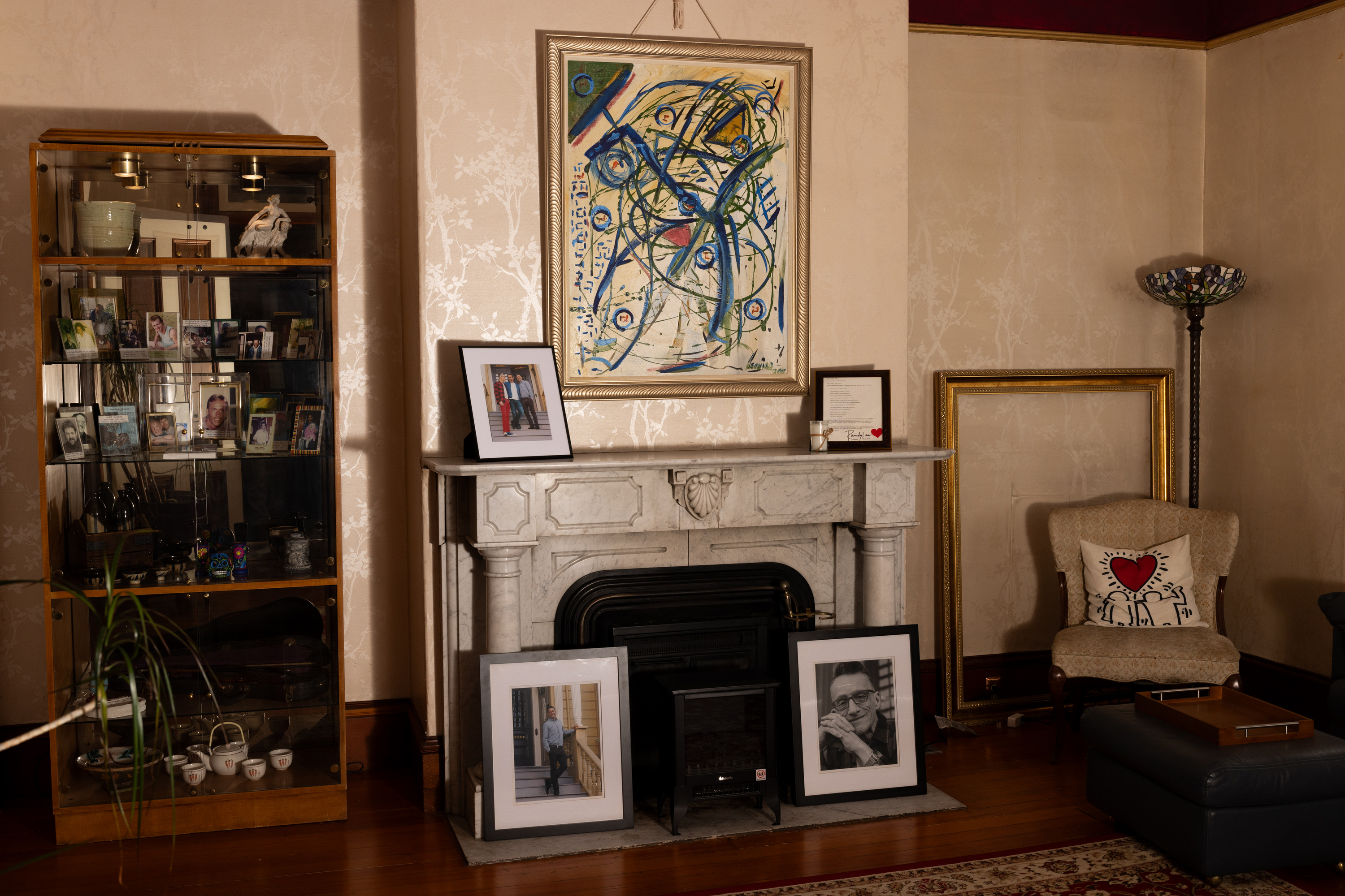 The image shows a room with a marble fireplace, abstract art above it, framed photos on the mantle, a display cabinet filled with pictures, and an armchair with a heart pillow.