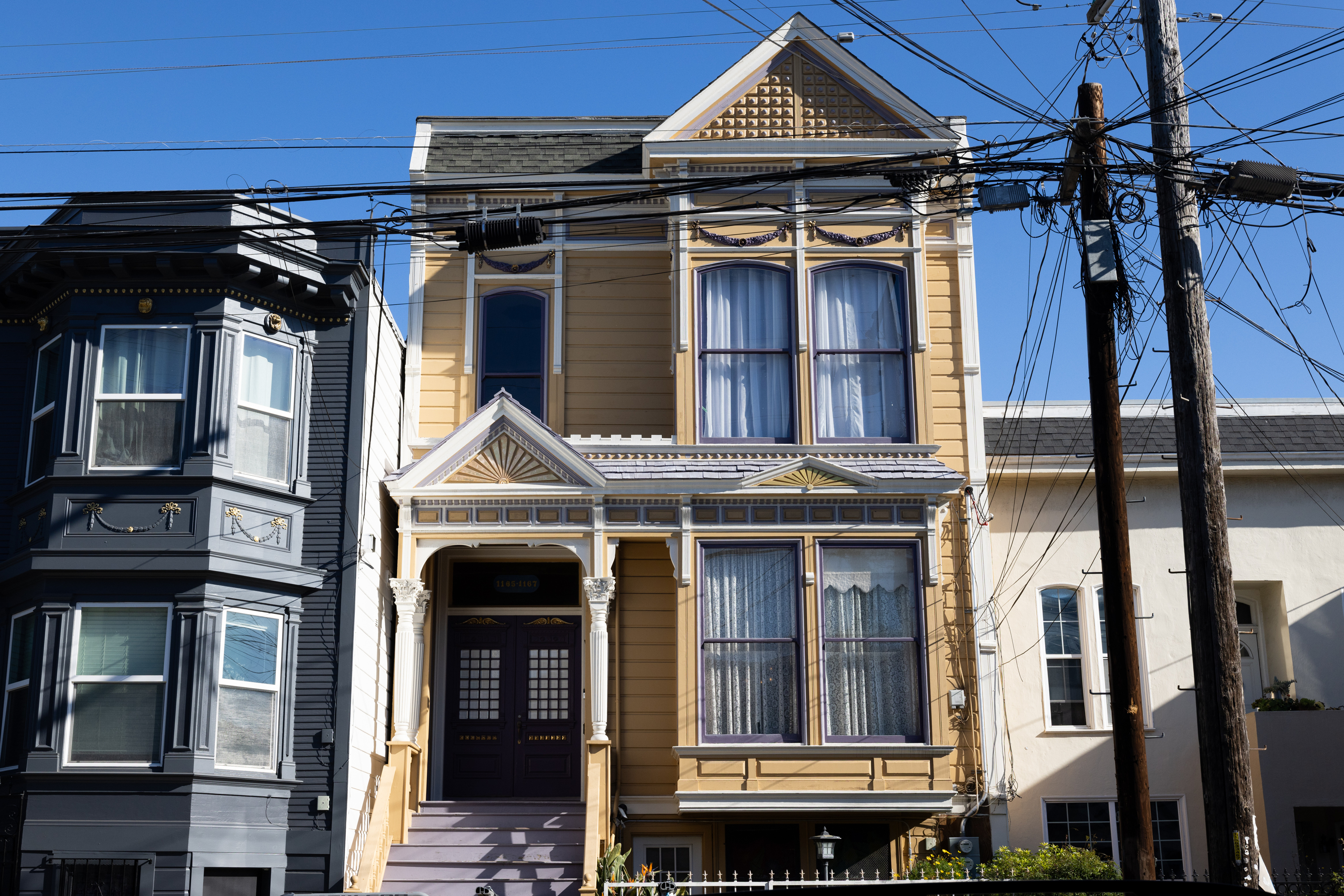 The image shows a yellow Victorian-style house with ornate decorations and tall windows. It is flanked by darker and lighter houses, with visible overhead wires.