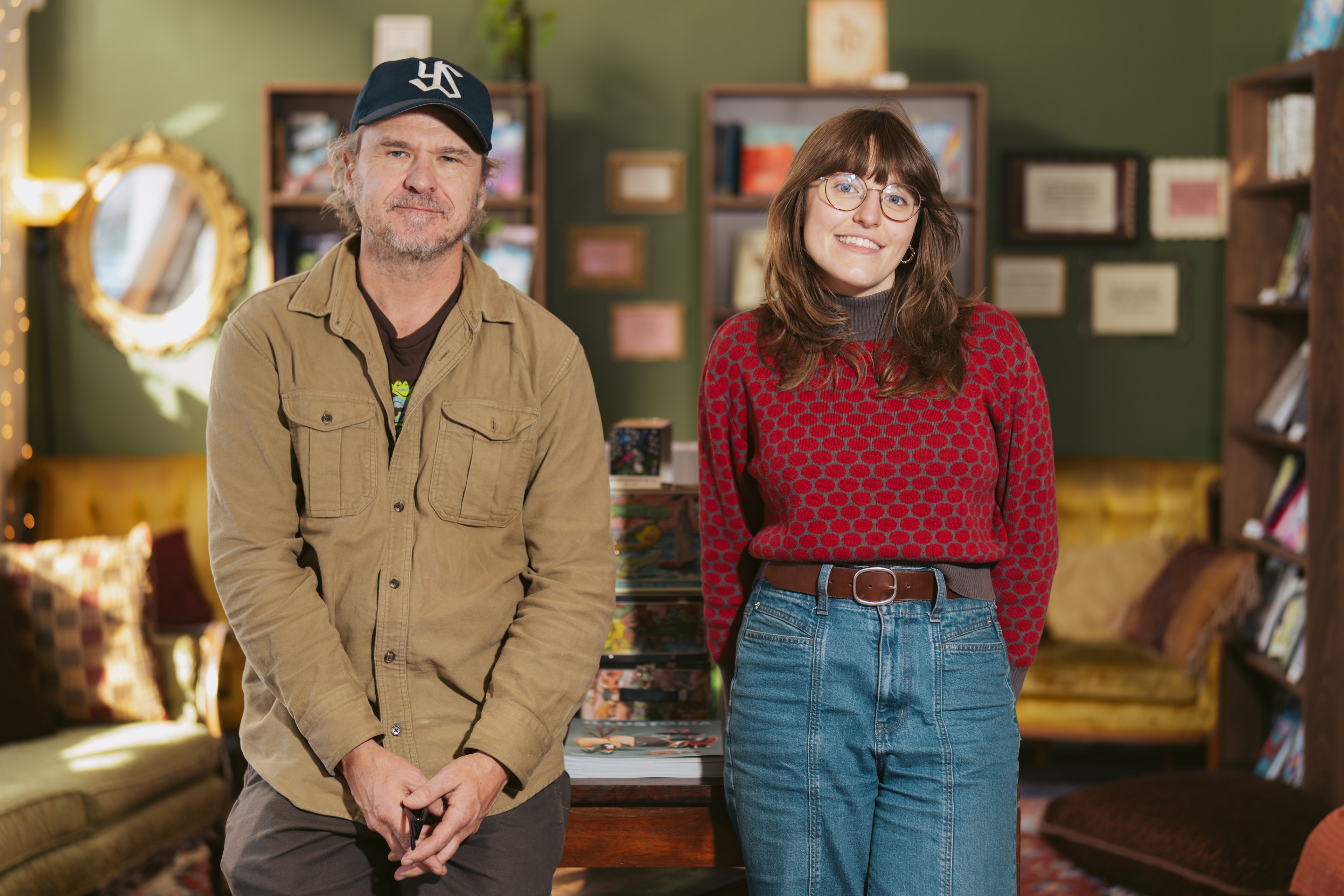 A man in a beige jacket and hat and a woman in a red sweater stand in a cozy room with bookshelves, a mirror, and comfortable seating in the background.