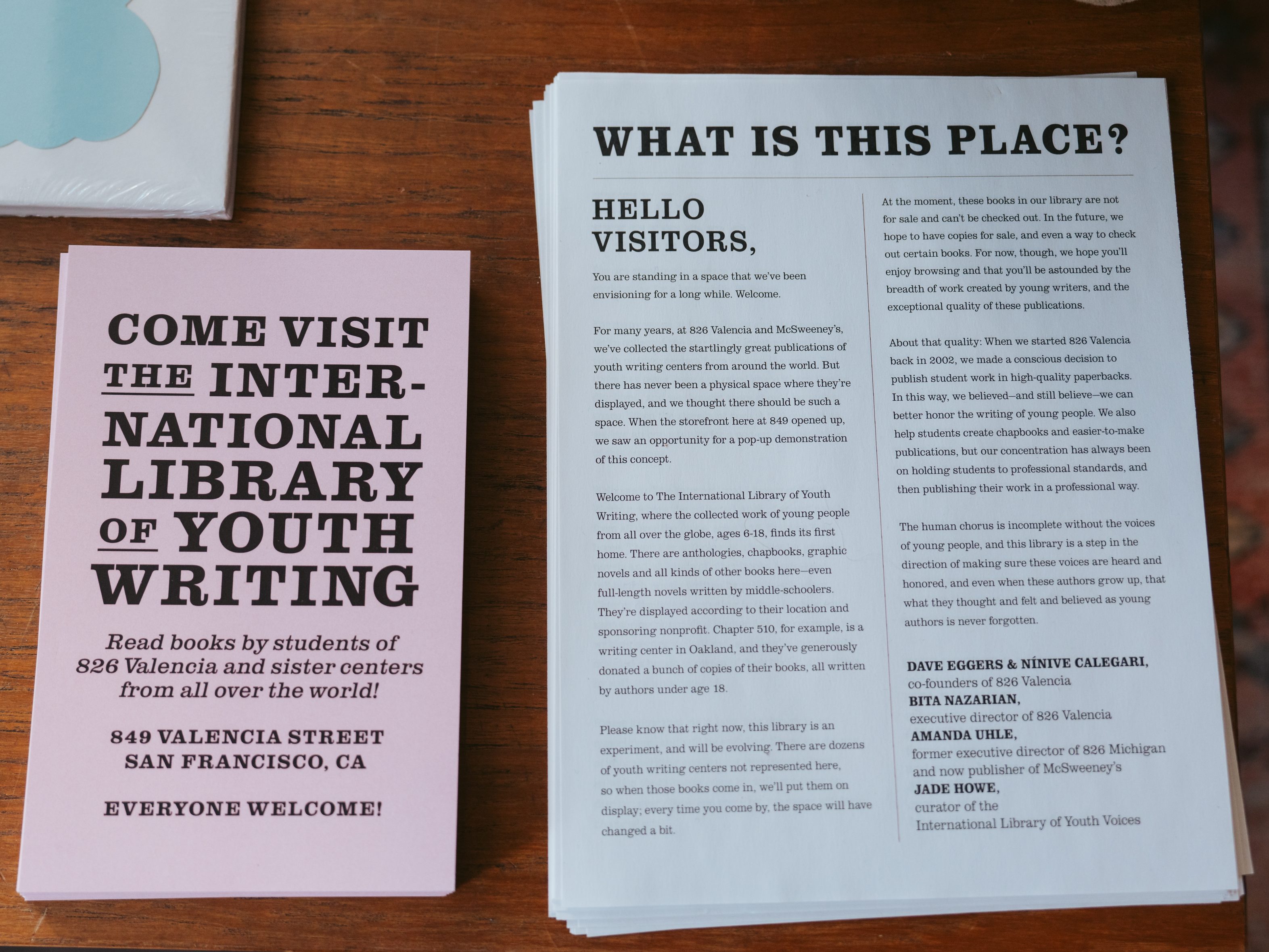 The image shows a pink flyer and a white informational card on a wooden surface, inviting people to visit the International Library of Youth Writing.