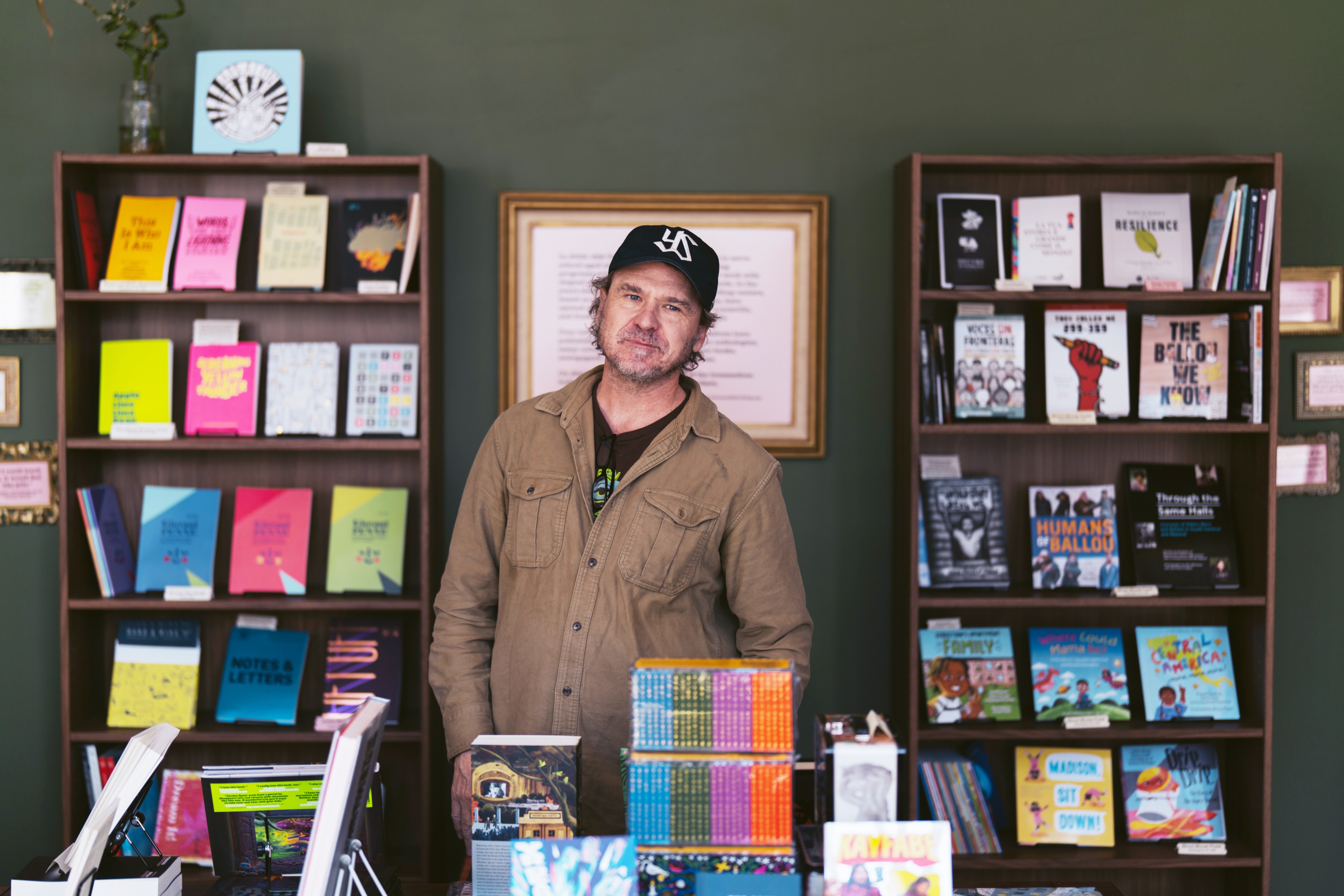 A man in a brown jacket and black cap stands in front of book-filled shelves. The books are colorful, and a framed picture is on the wall behind him.