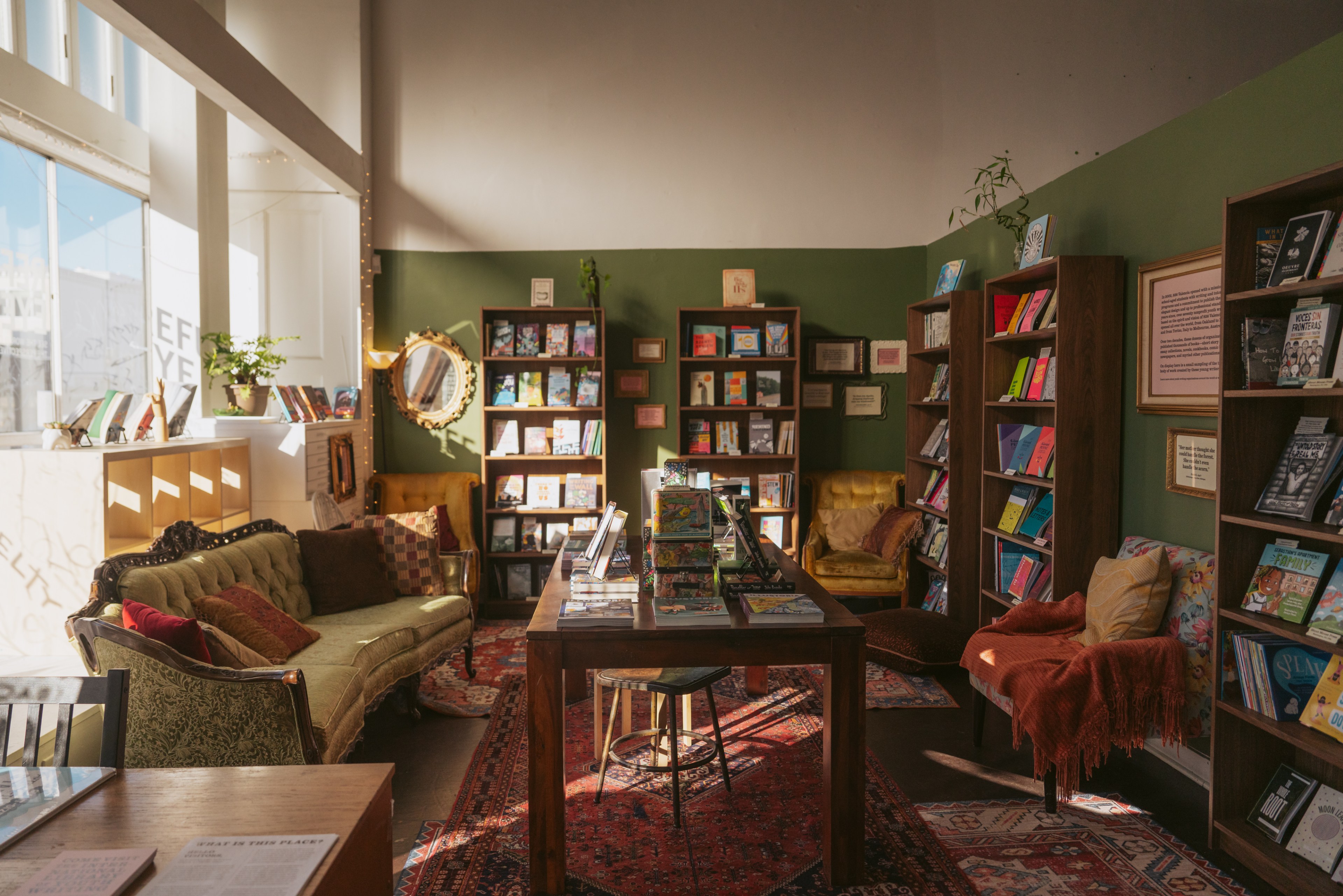 A cozy bookstore with green walls, filled with bookshelves. There are plush sofas and chairs, a central wooden table, warm lighting, and decorative plants.