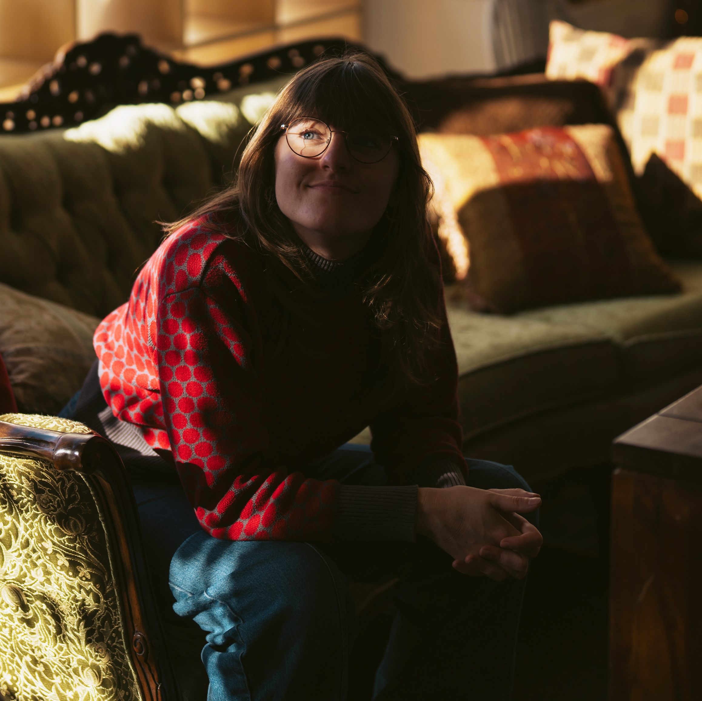 A person with long hair and glasses sits on a sofa in a warmly lit room. They're wearing a red-patterned sweater and have a relaxed expression.