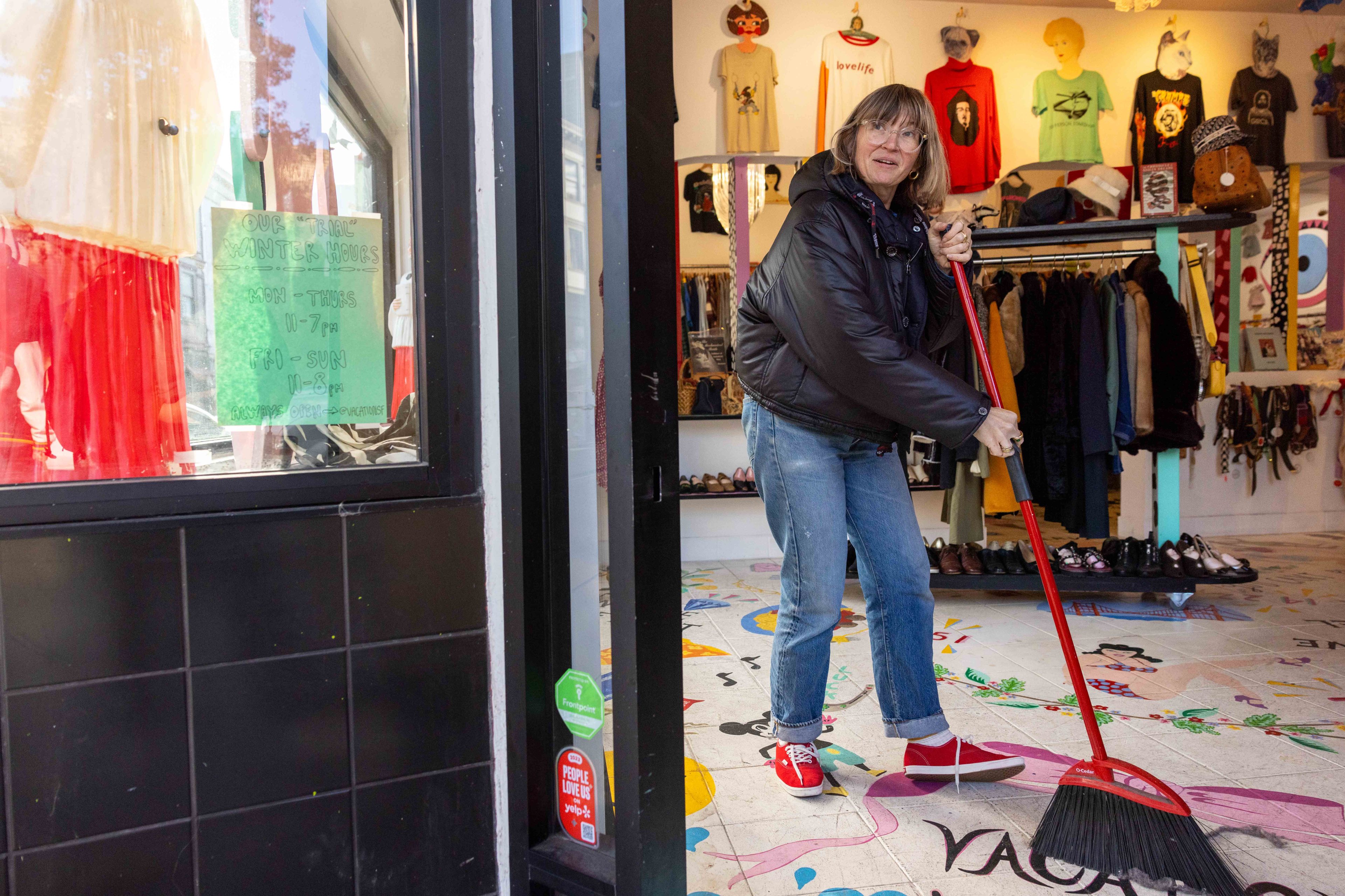 A person in a black jacket and jeans sweeps the floor in a clothing store filled with colorful shirts on display. A sign is visible on the shop window.