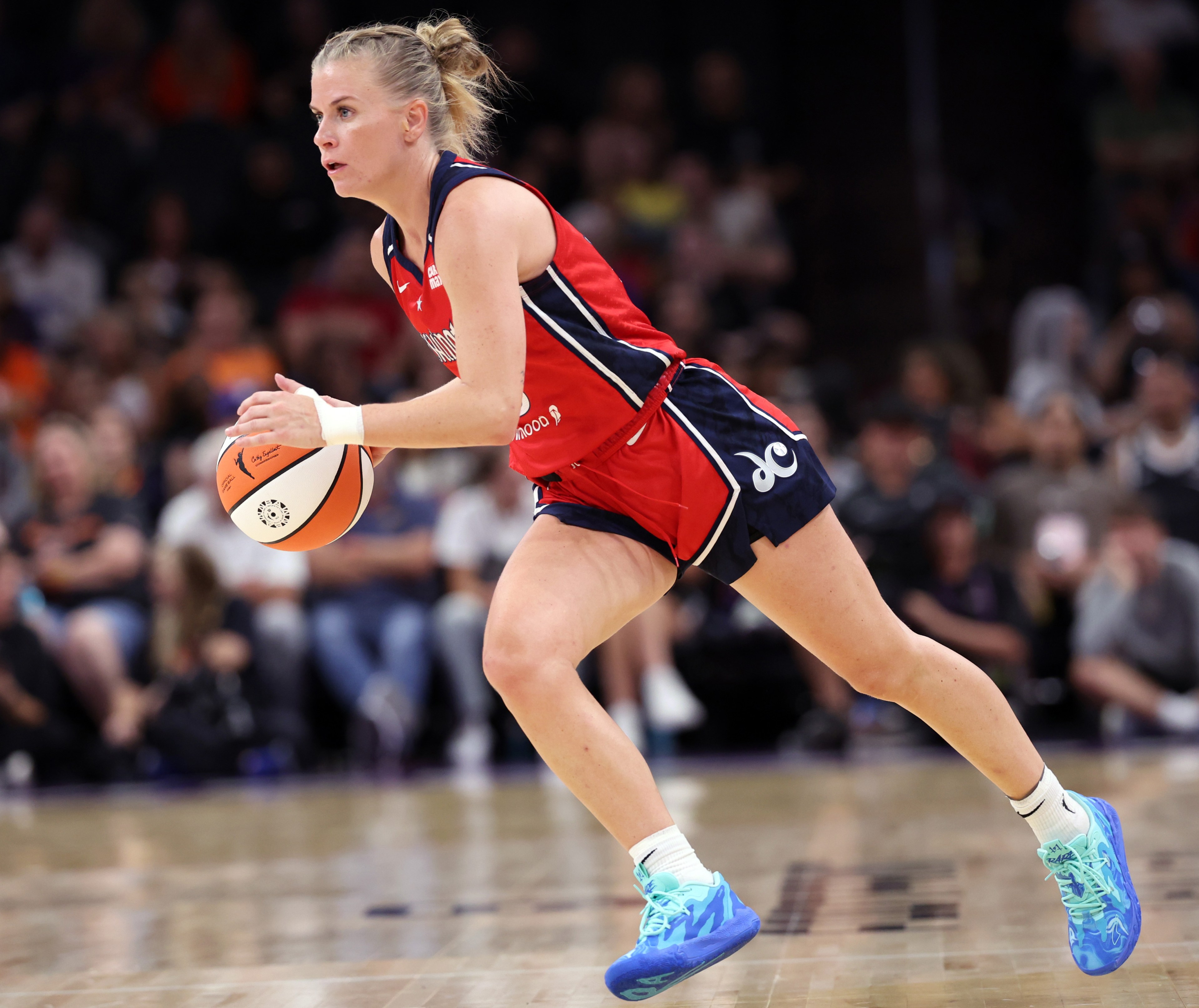 A basketball player in a red uniform is dribbling a ball on the court, focused and in motion, with an audience in the blurred background.