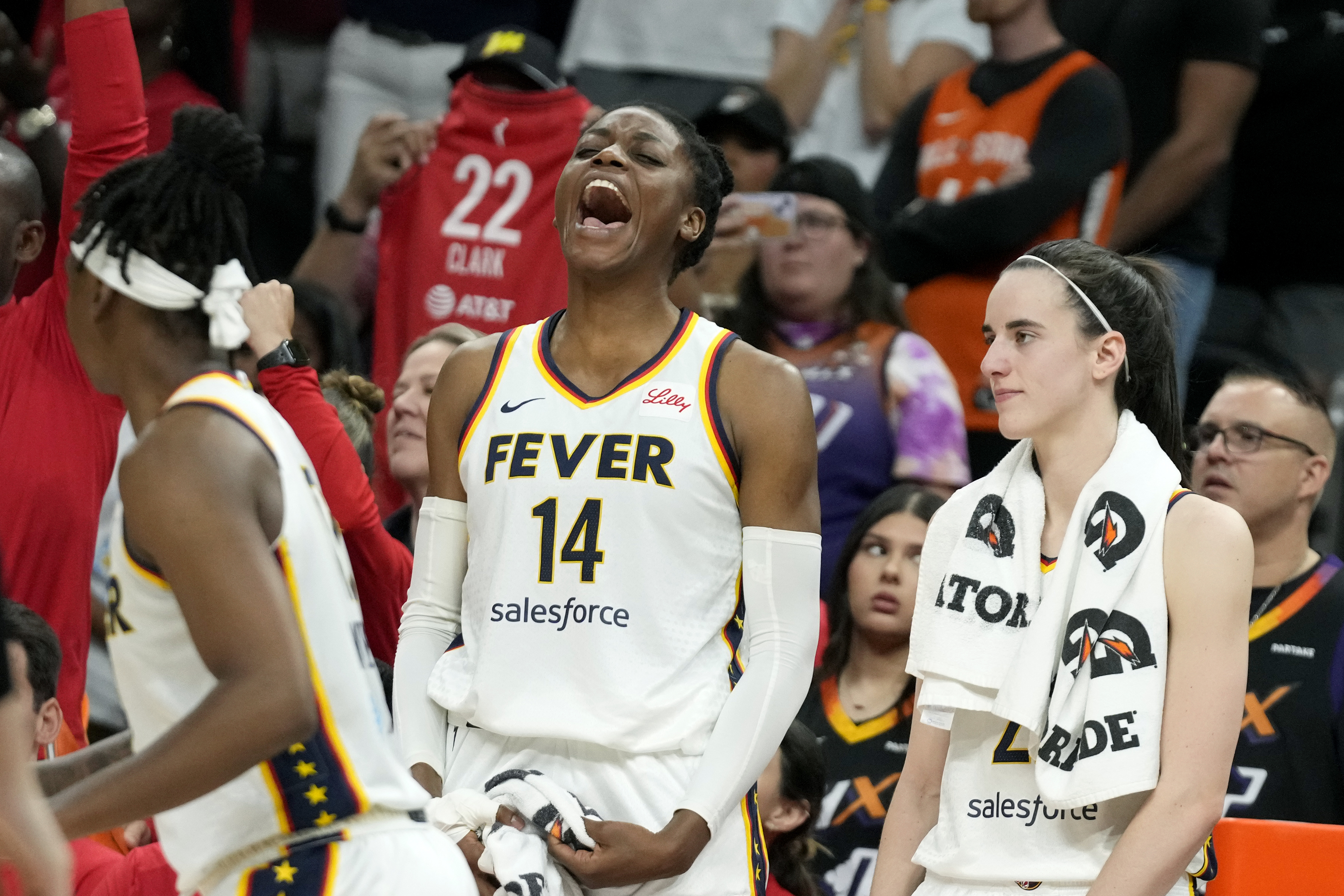 A basketball player wearing a &quot;Fever&quot; jersey shouts excitedly on the court, surrounded by teammates and fans in the background, creating an energetic atmosphere.