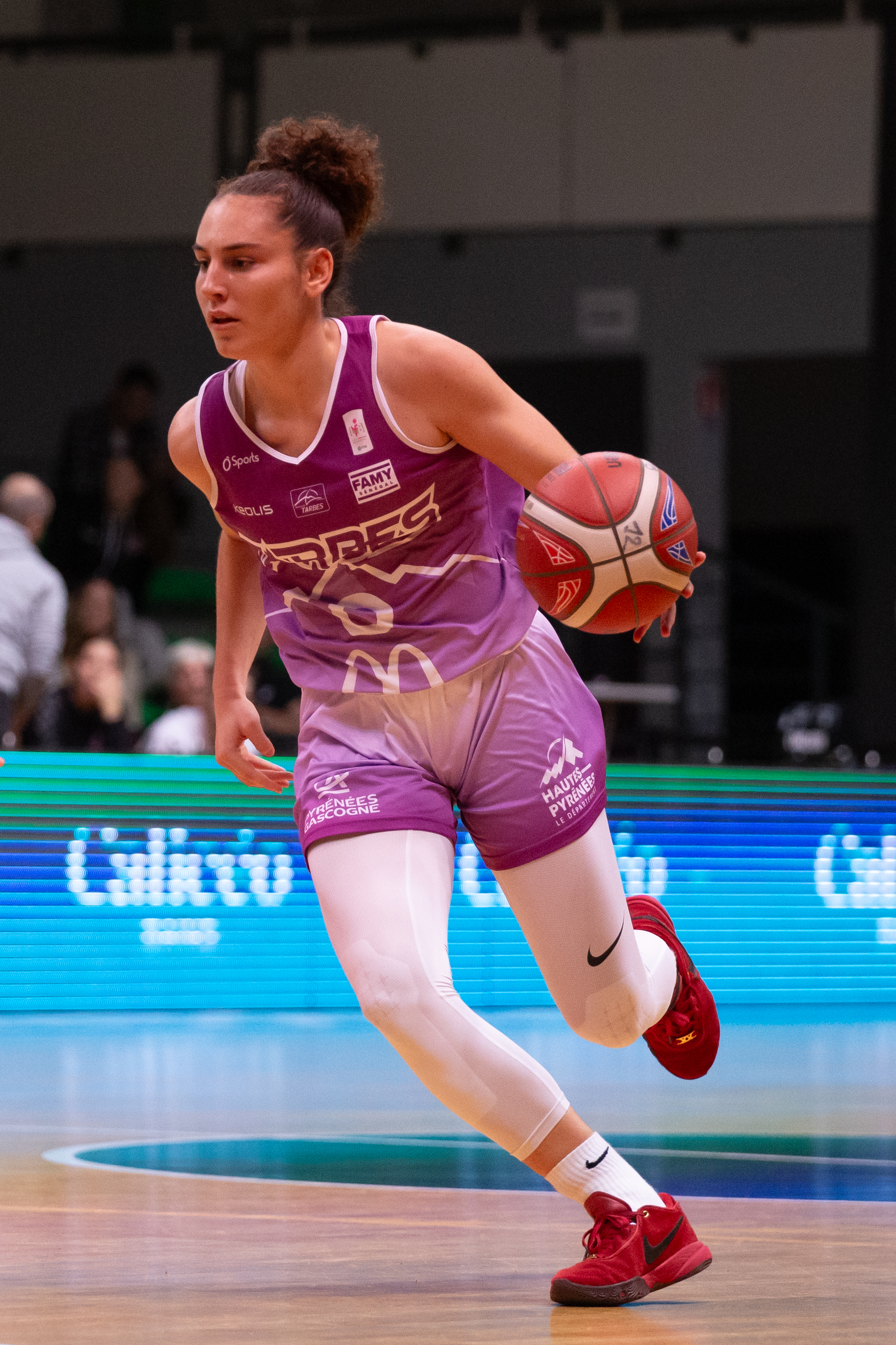 A basketball player in a purple jersey and shorts dribbles a basketball. She has curly hair tied up, wears white leggings, and red sneakers on a court.