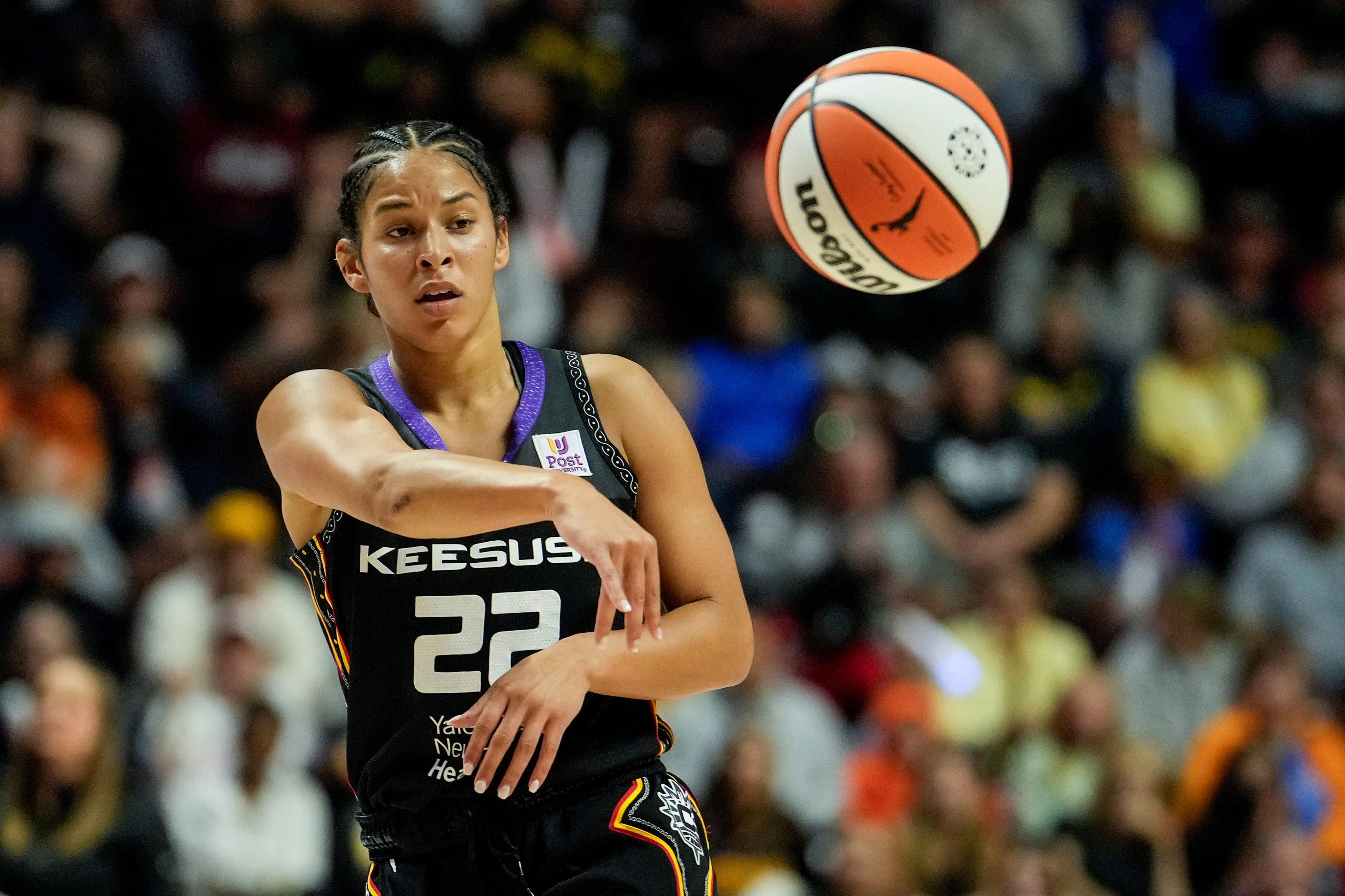 A basketball player in a black jersey with number 22 is focused as she throws a basketball. The crowd in the background is blurred.