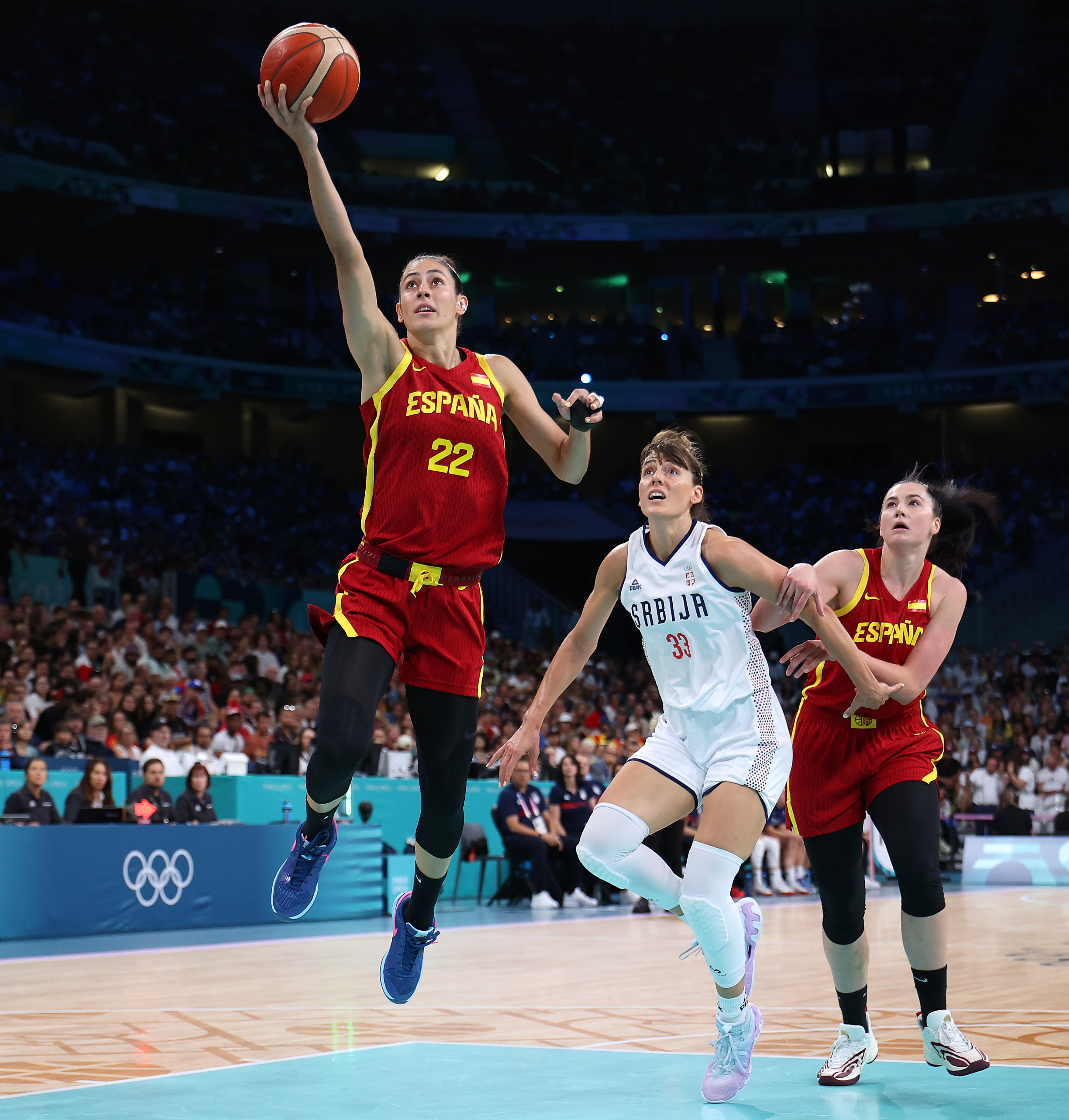 A basketball player in a red &quot;España&quot; jersey is leaping to make a layup. Two other players, one in a white &quot;Srbija&quot; jersey and another in red, are nearby.