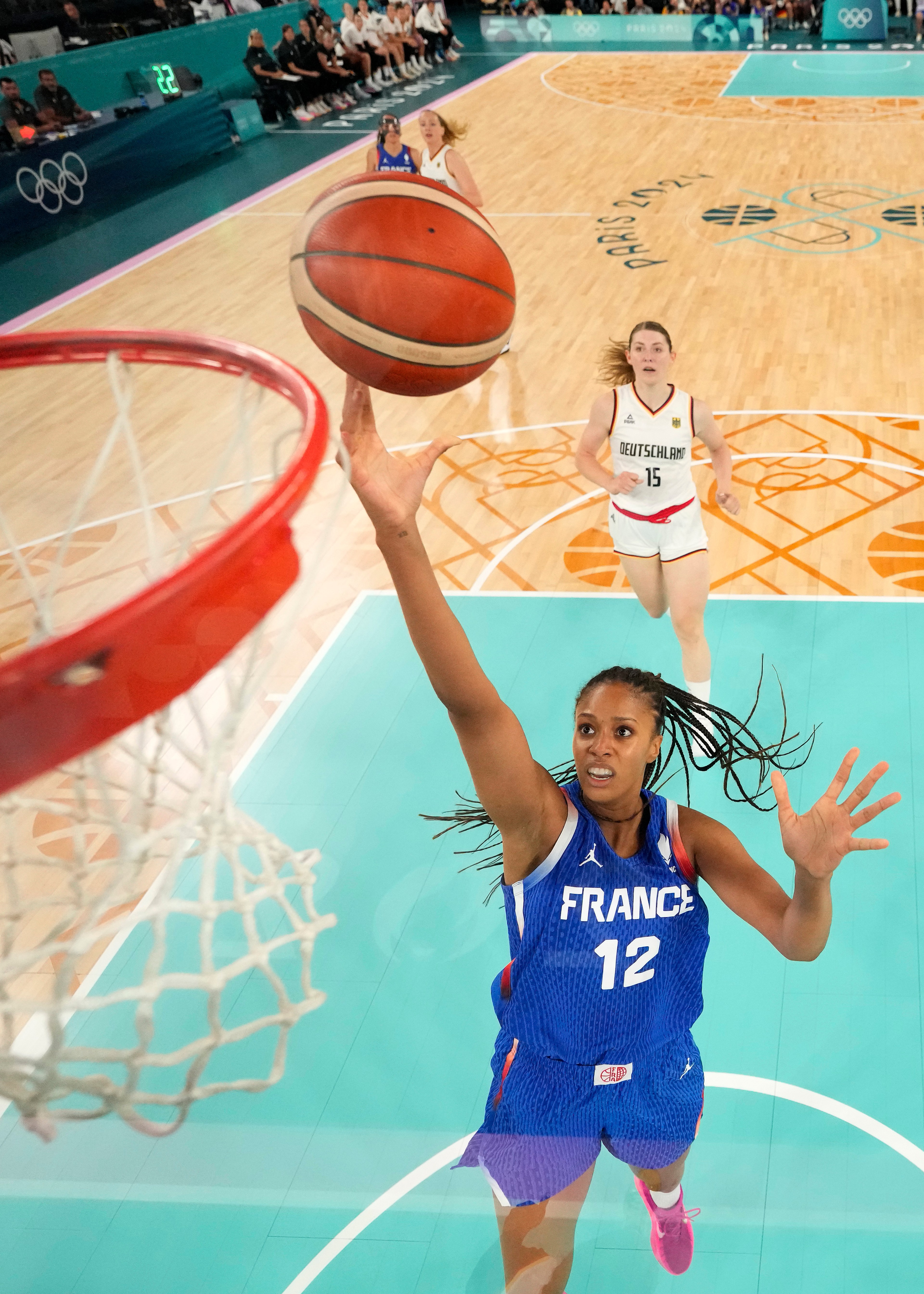 A basketball player in a France jersey is leaping to score, with a Germany player behind her on a brightly colored court, viewed from above the hoop.