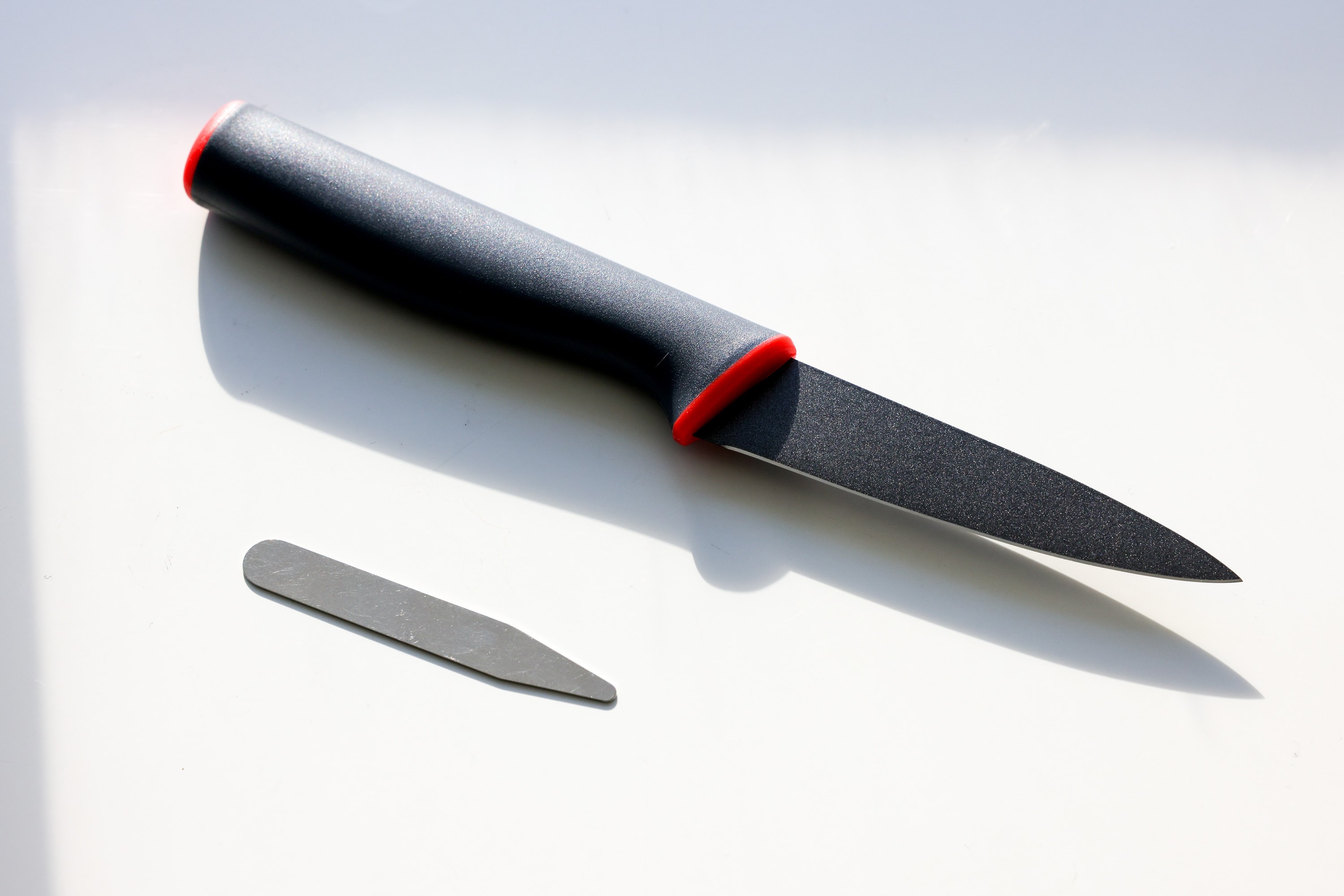 A black knife with a red accent on the handle is placed on a light surface, casting a shadow. Nearby, a small, flat metal collar stay is also in the image.