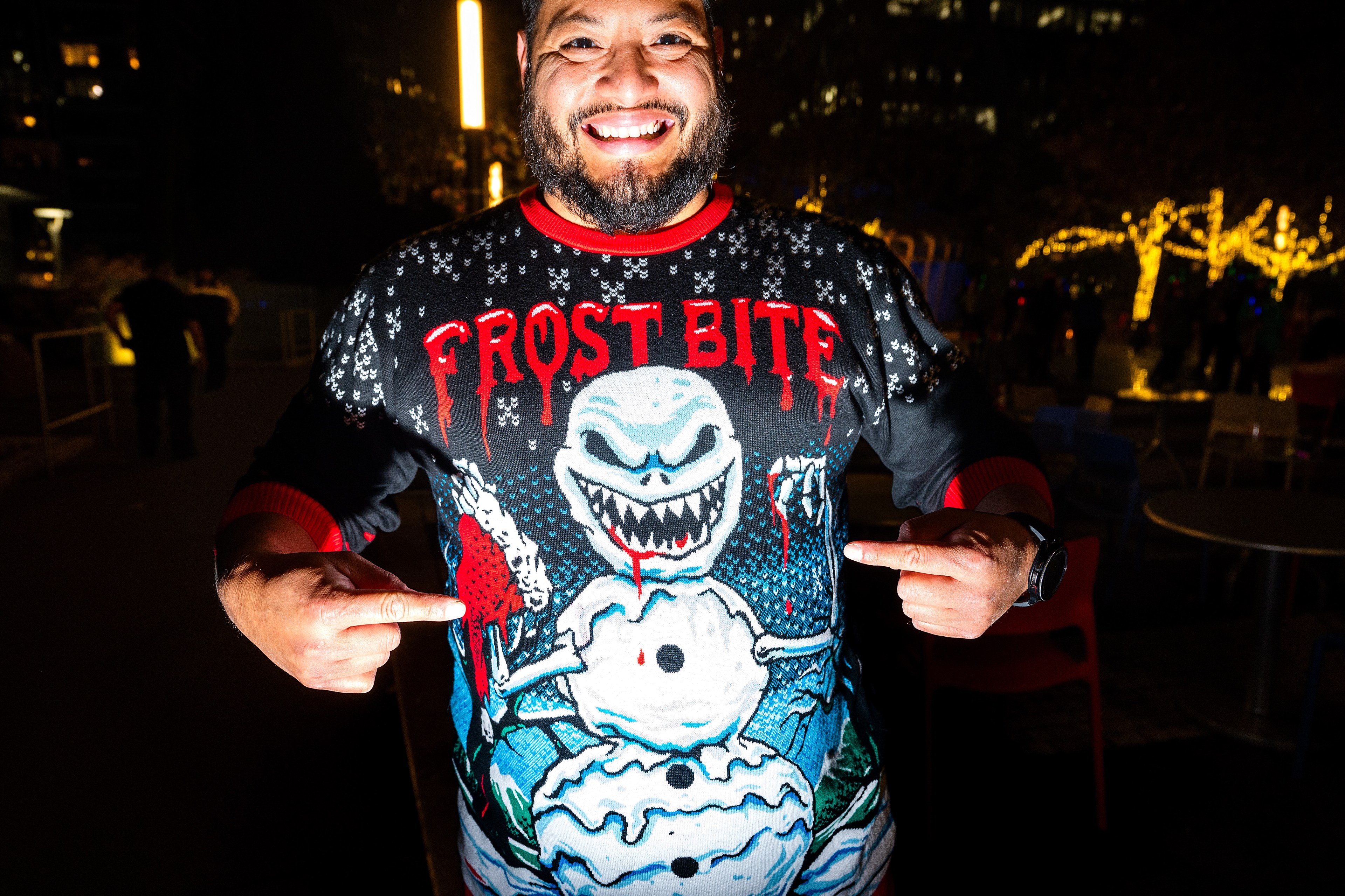 A man is smiling and pointing at his sweater, which features a menacing snowman with jagged teeth, blood accents, and the text "FROST BITE."