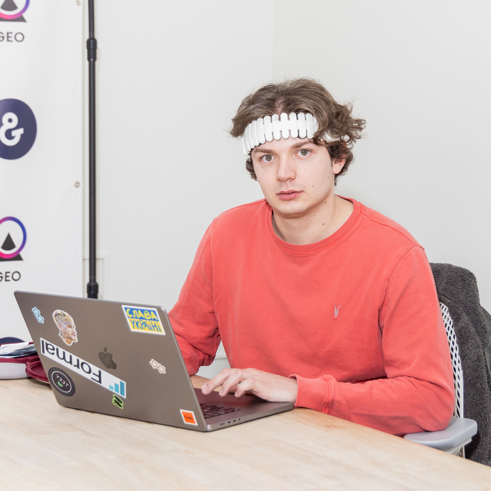 A person in a red shirt and a unique headband is sitting at a table using a laptop with various stickers. The background features simple white walls and logos.