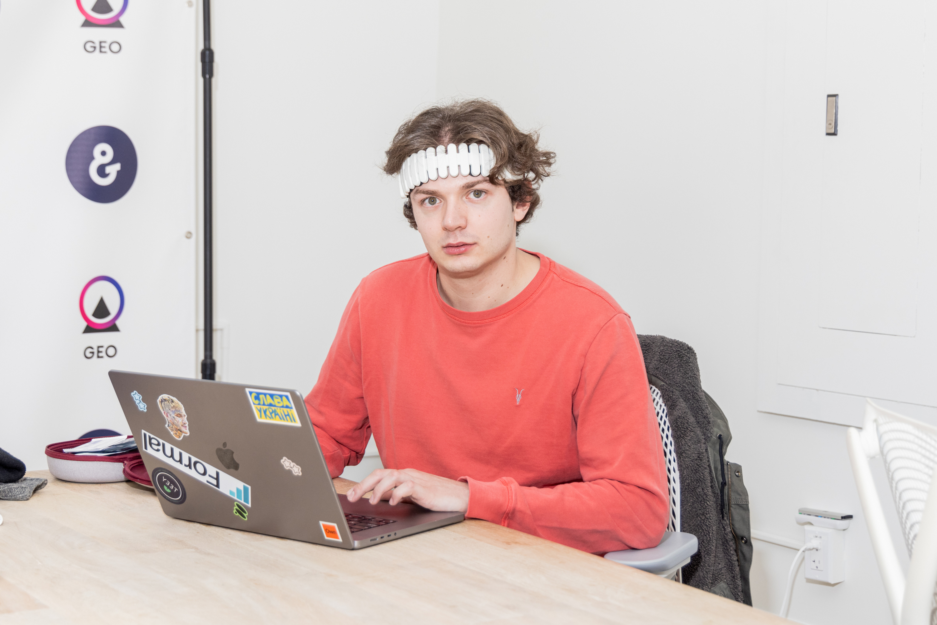 A person in a red shirt and a unique headband is sitting at a table using a laptop with various stickers. The background features simple white walls and logos.