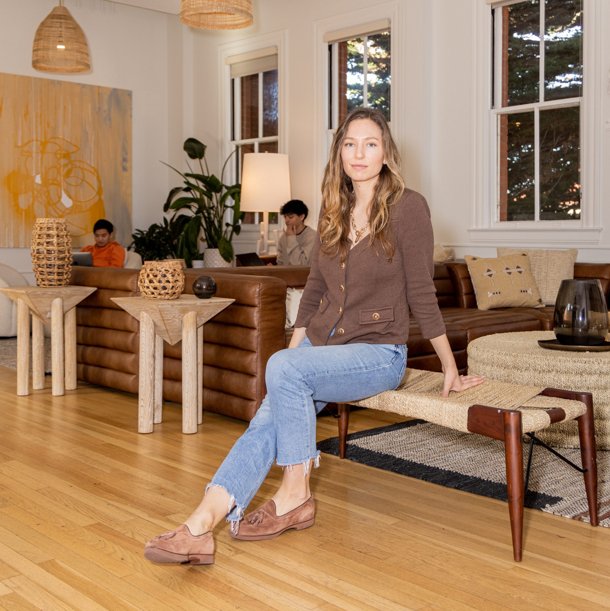 A person sits on a woven bench in a cozy living room with brown sofas, wicker tables, and abstract art. Two others work on laptops, surrounded by plants and soft lighting.