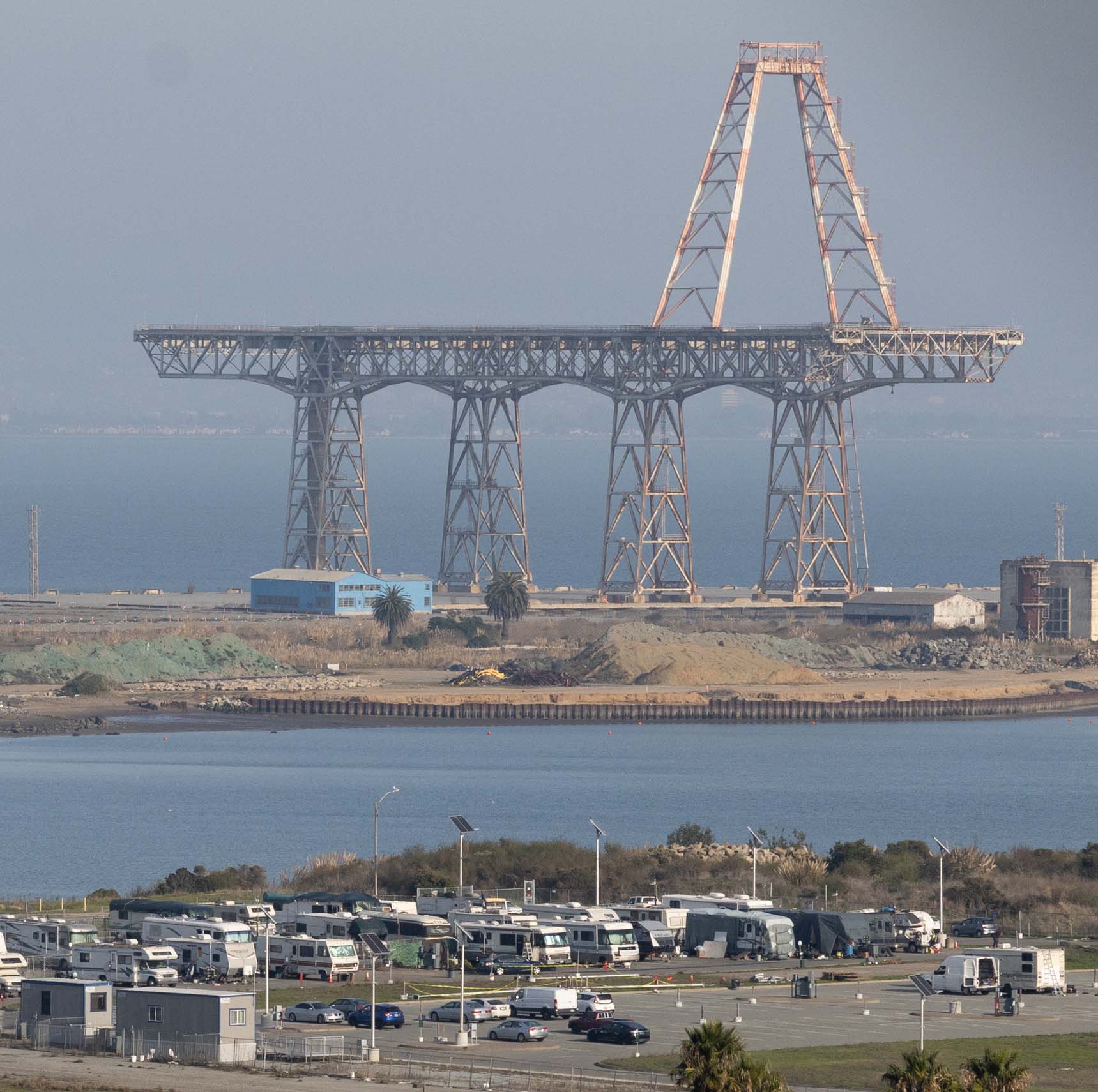 The image shows a large, incomplete steel bridge structure by a body of water. In the foreground, there are parked RVs and cars on a paved area.