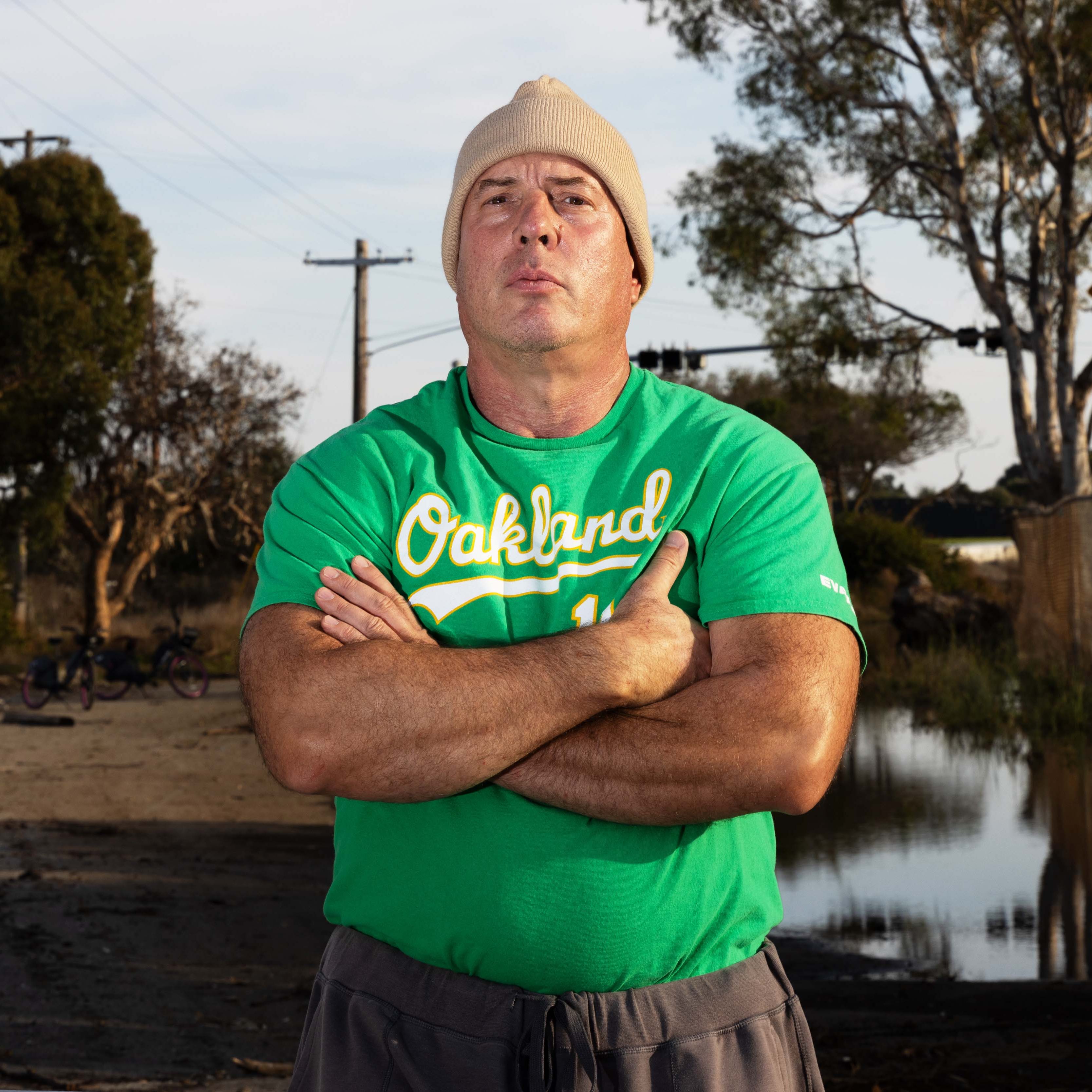 A person in a green &quot;Oakland&quot; shirt and beige beanie crosses their arms confidently. The backdrop features trees, power lines, and a calm water reflection.