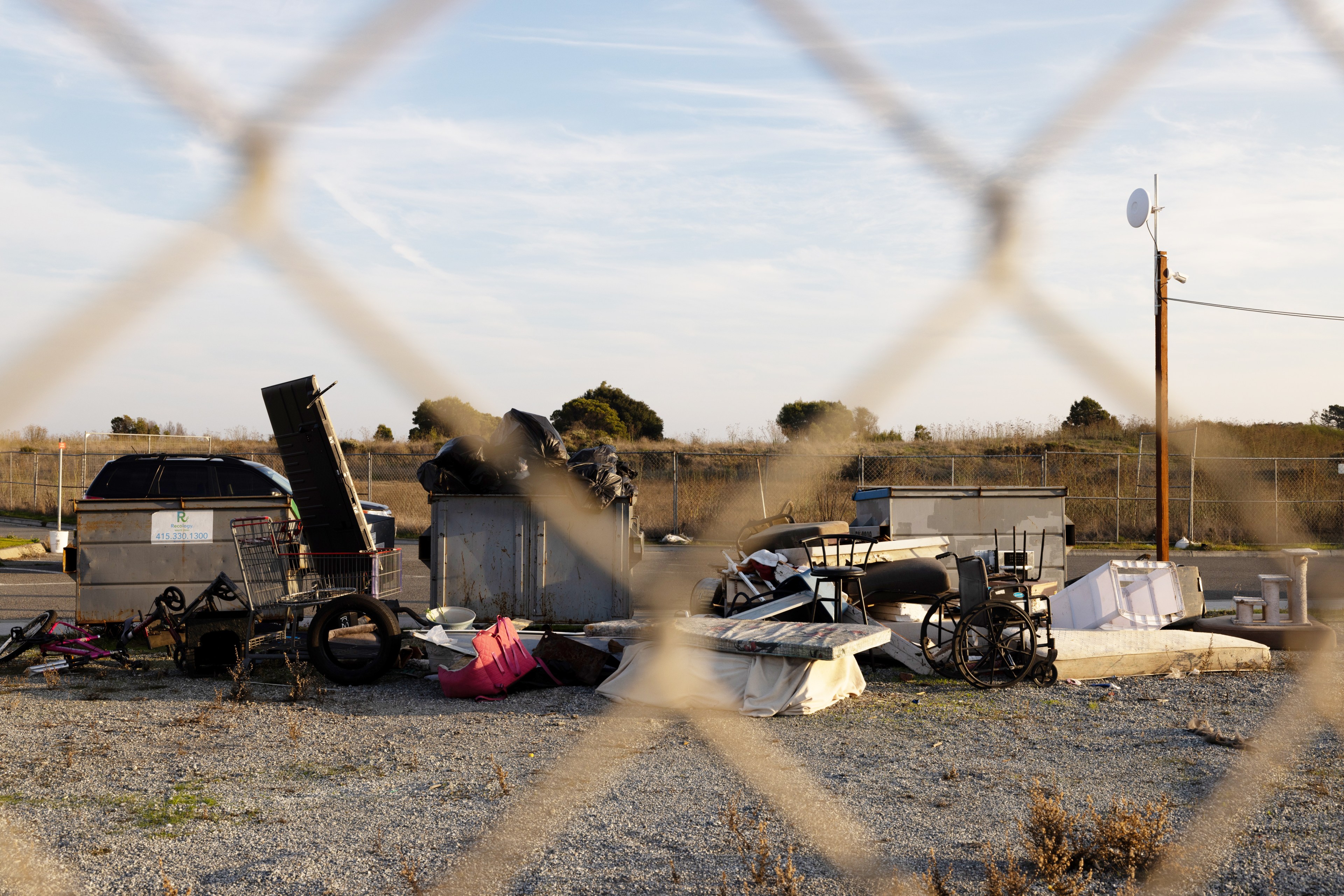 A fenced-off area contains discarded items like a wheelchair, shopping cart, and debris under a clear sky with distant trees.