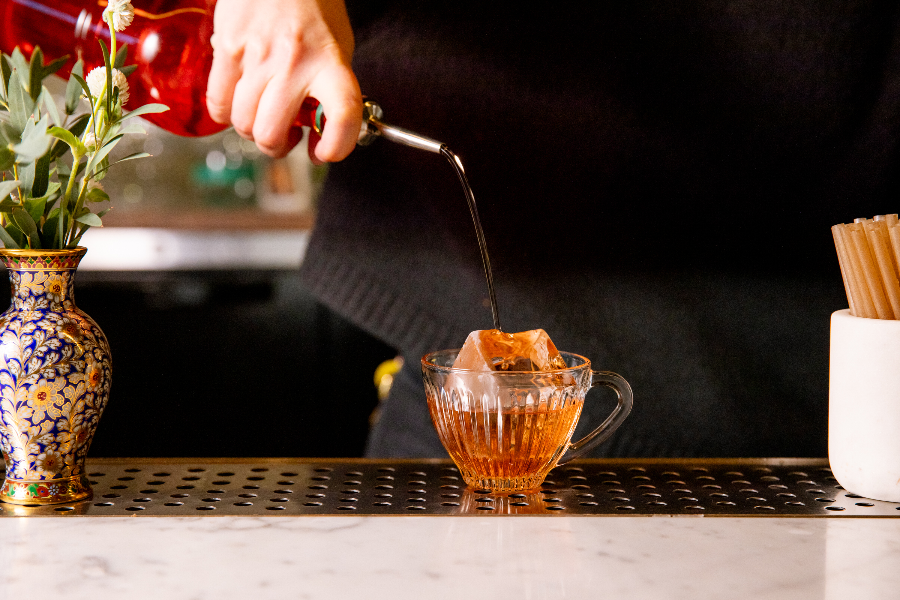 A hand pours dark liquid from a decanter into a glass cup with ice. A floral vase and straws in a container are on a marble counter nearby.