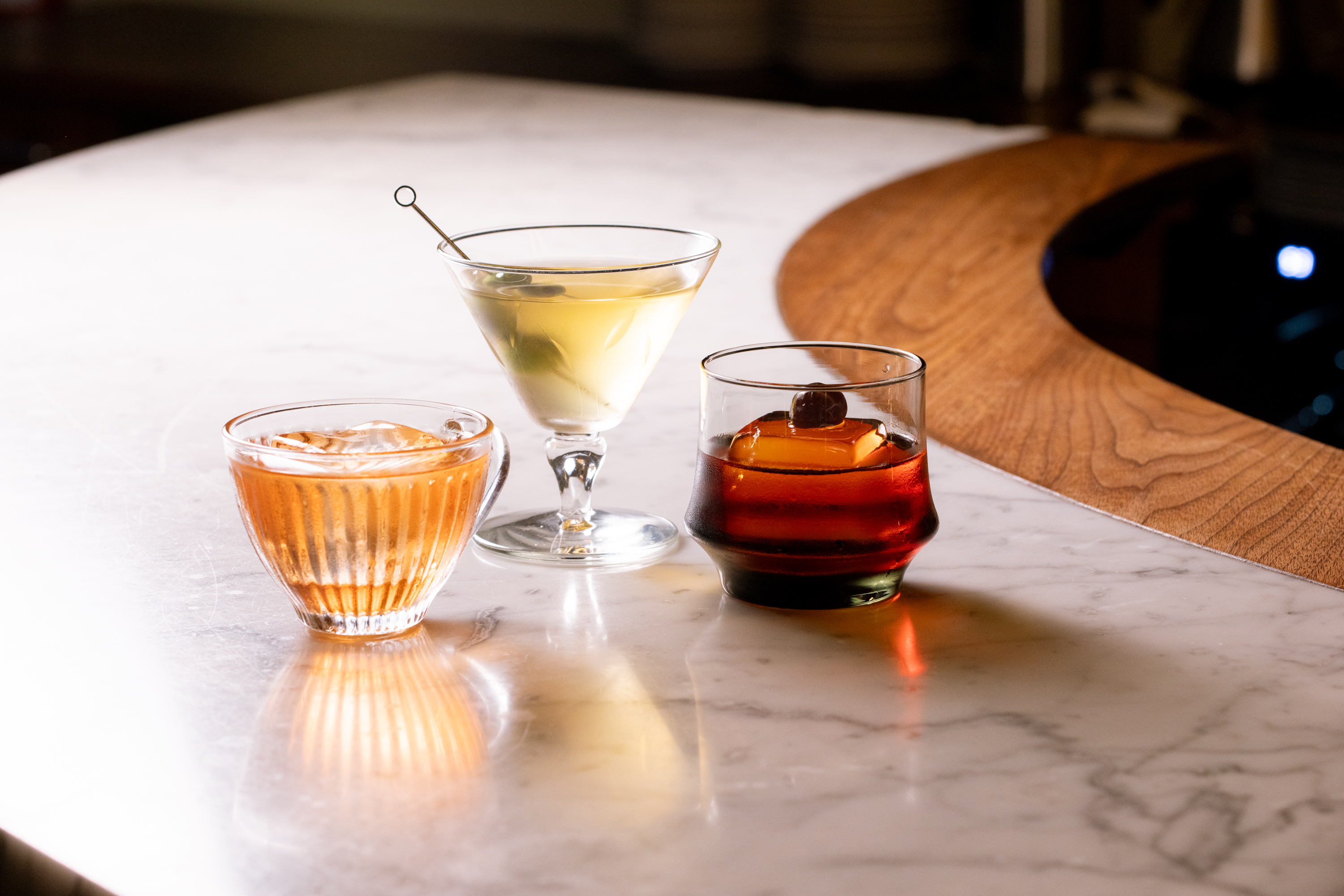 Three cocktails sit on a marble bar. A ridged glass holds an amber drink with ice, a martini glass contains a clear drink with an olive, and a tumbler has a dark drink with a cherry.