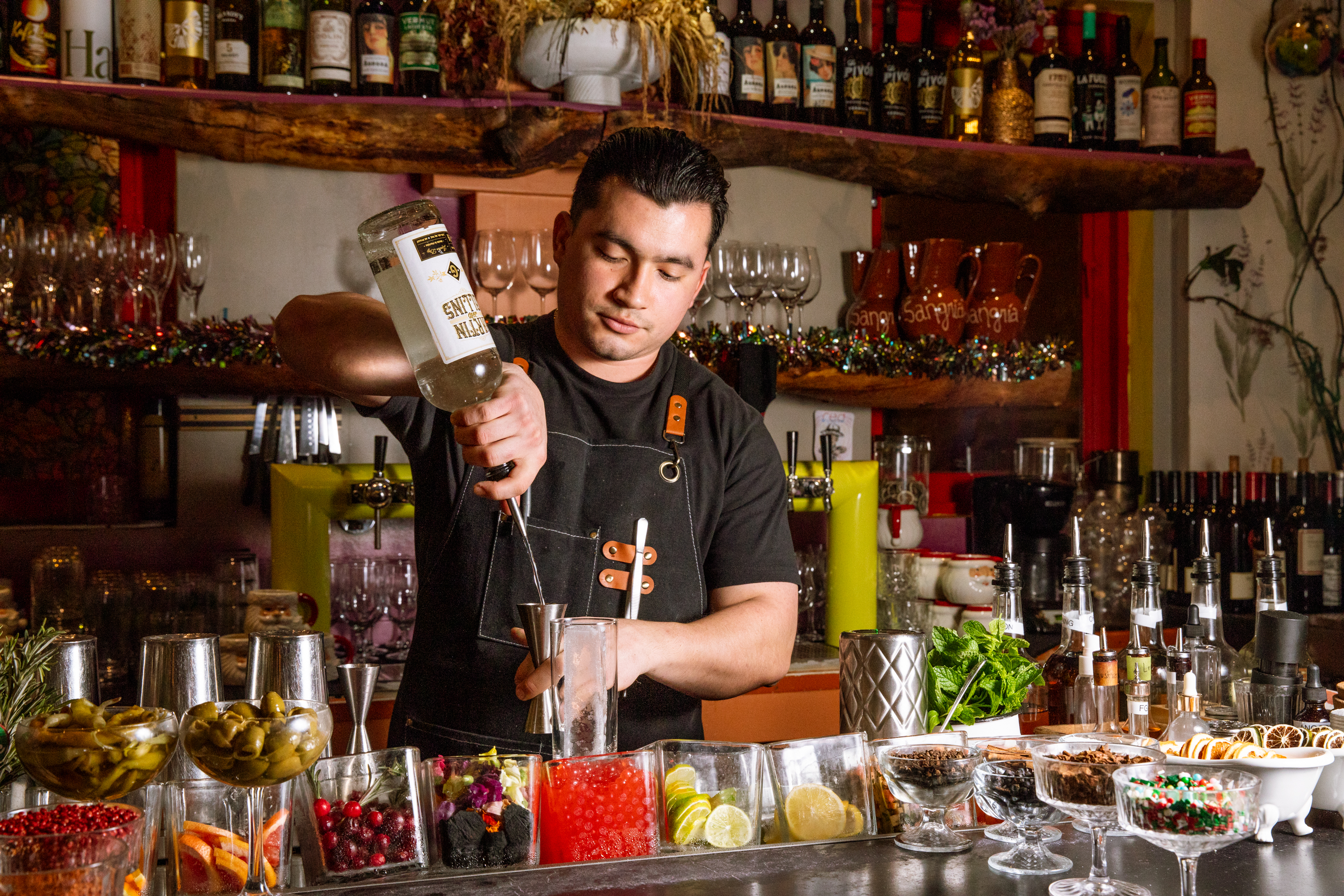 A bartender pours liquid into a shaker over a bar lined with various garnishes. Shelves of bottles and glasses are visible in the background.