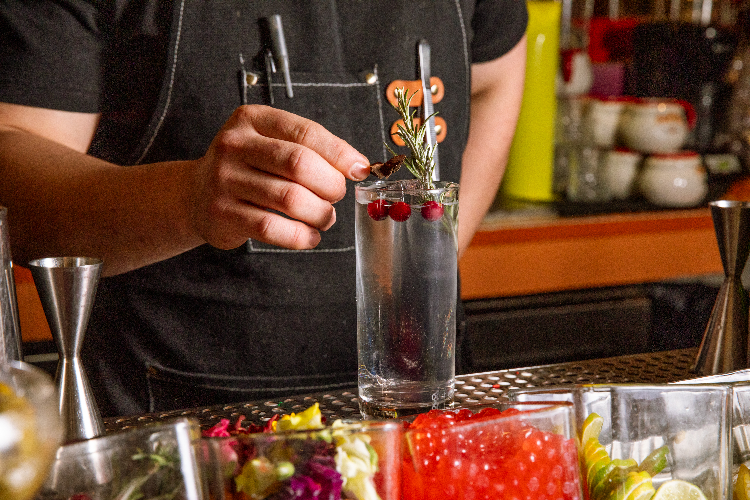 A bartender garnishes a cocktail with a sprig of rosemary and cranberries in a tall glass. Various colorful ingredients are on the bar counter.