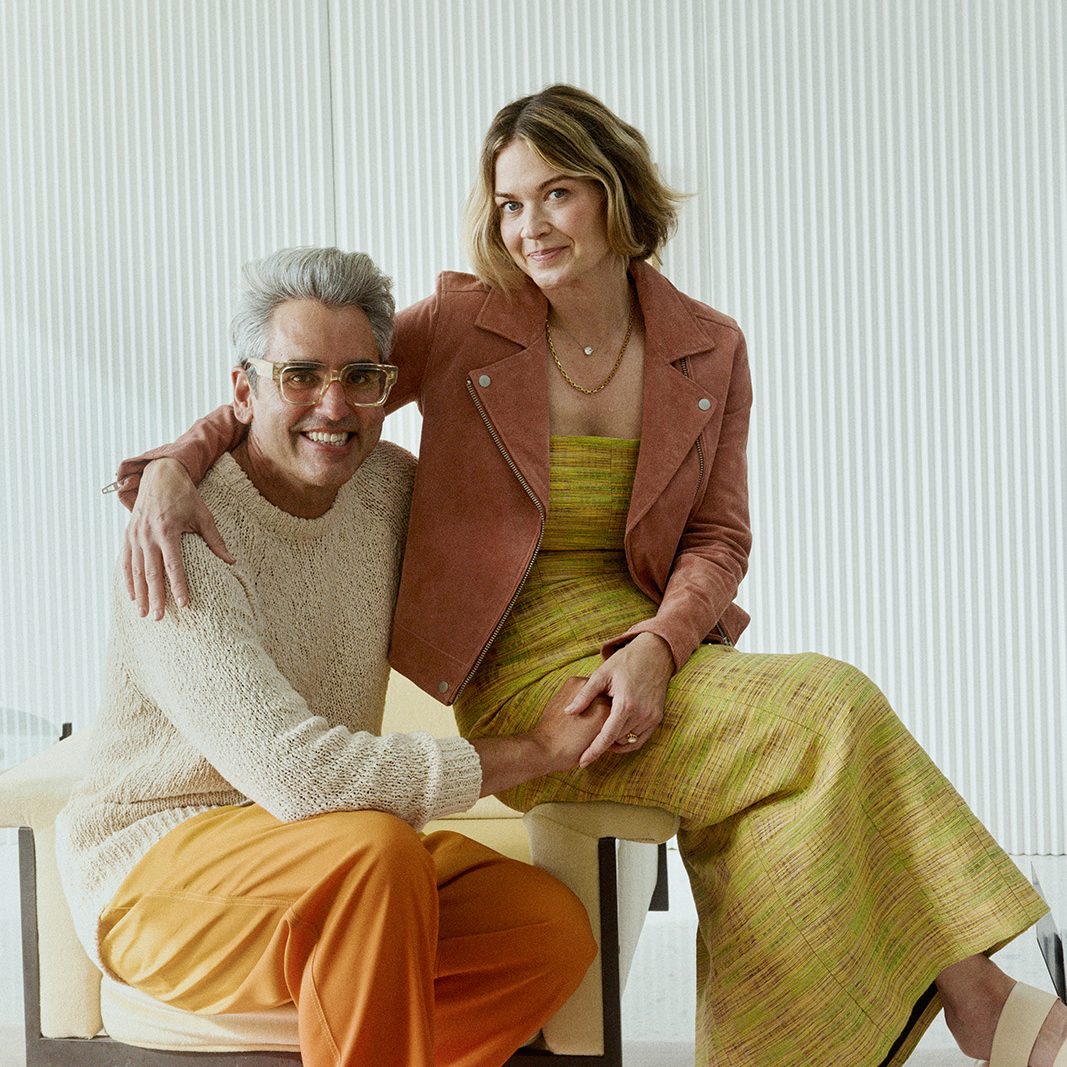 A smiling couple sits together. The man wears glasses, an off-white sweater, and orange pants. The woman is in a striped dress with a brown jacket. They hold hands.