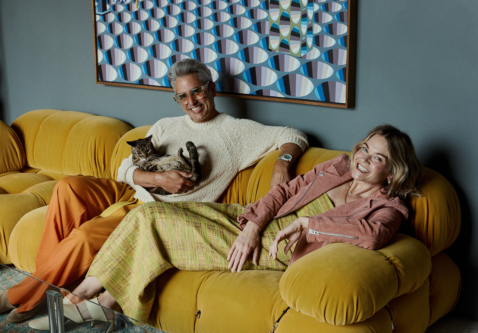 A man and woman sit on a yellow sofa, smiling. The man is holding a cat. Behind them hangs a colorful geometric painting. They appear relaxed and happy.