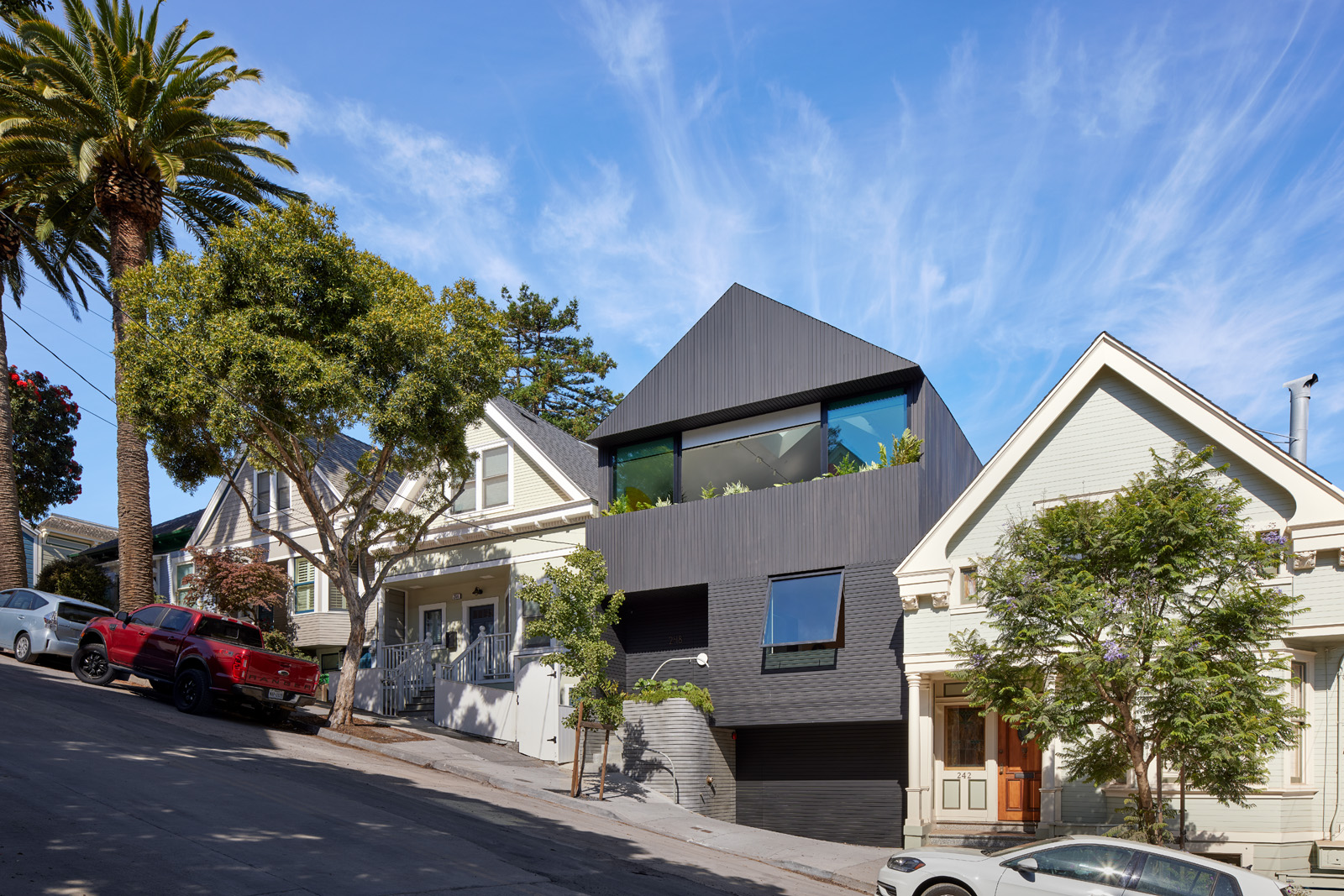 A modern black house with large windows stands on a sloped street, flanked by traditional white homes and lush trees under a clear blue sky.