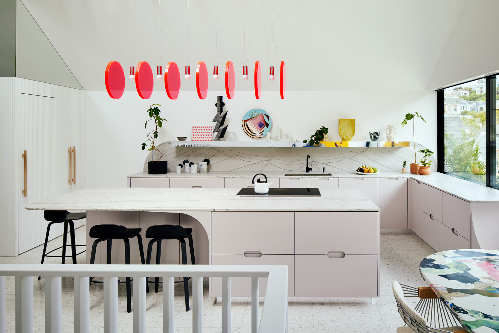 The image shows a modern kitchen with pastel cabinets, a marble island, black stools, unique red pendant lights, plants, and art on the shelves. A large window offers natural light.