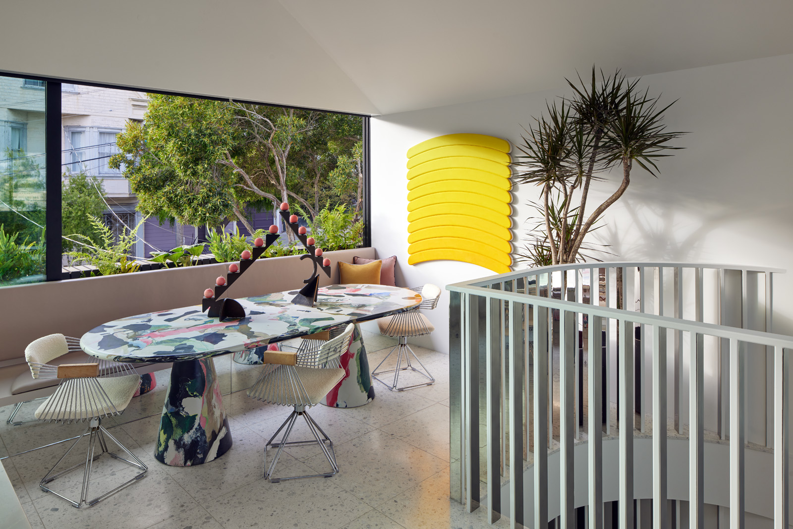 A modern room features a colorful marble table with wire chairs, large window view, yellow wall art, spiral staircase, and indoor plants.