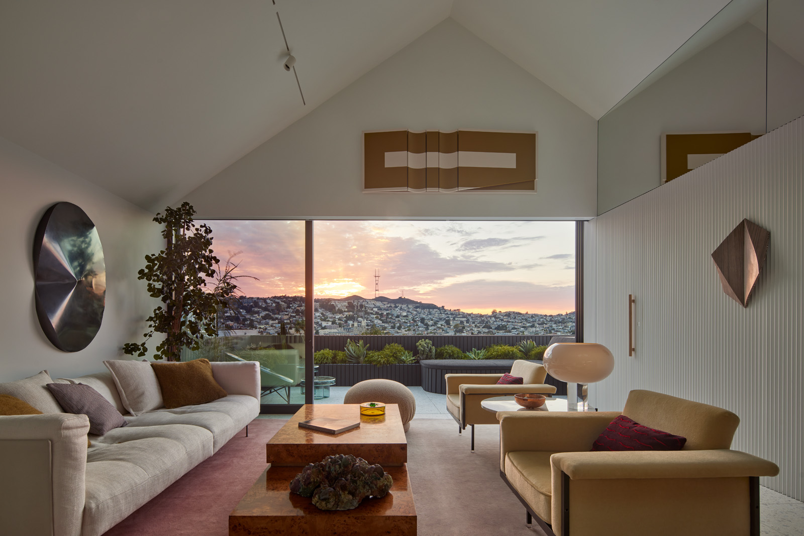 A modern living room features a large window with a sunset view, a white sofa, armchairs, abstract art, and a low table with decorative items. A potted plant stands nearby.