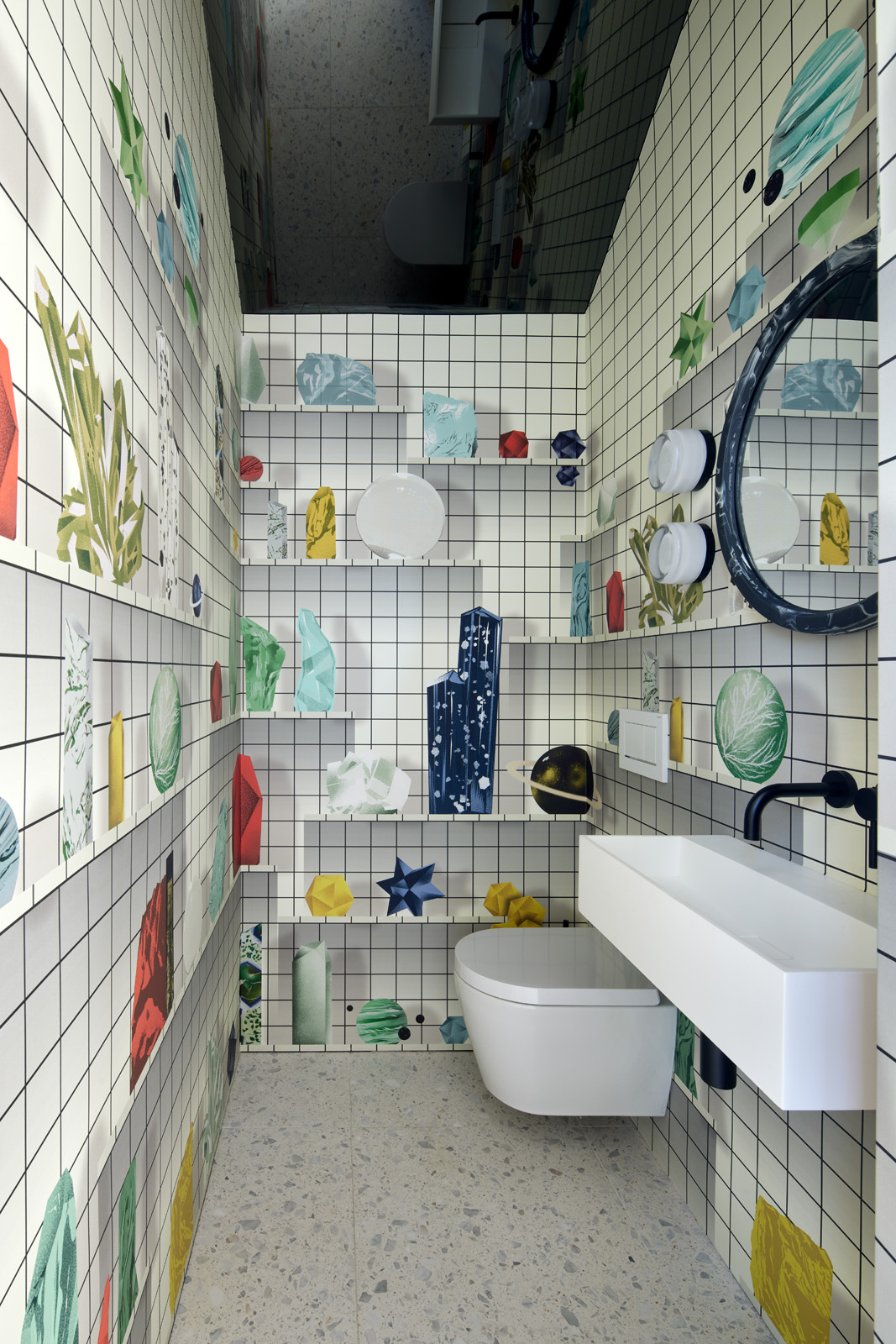 A small bathroom with grid-patterned walls, decorated with colorful geometric art. It features a wall-mounted toilet, a sleek white sink, and a circular mirror.