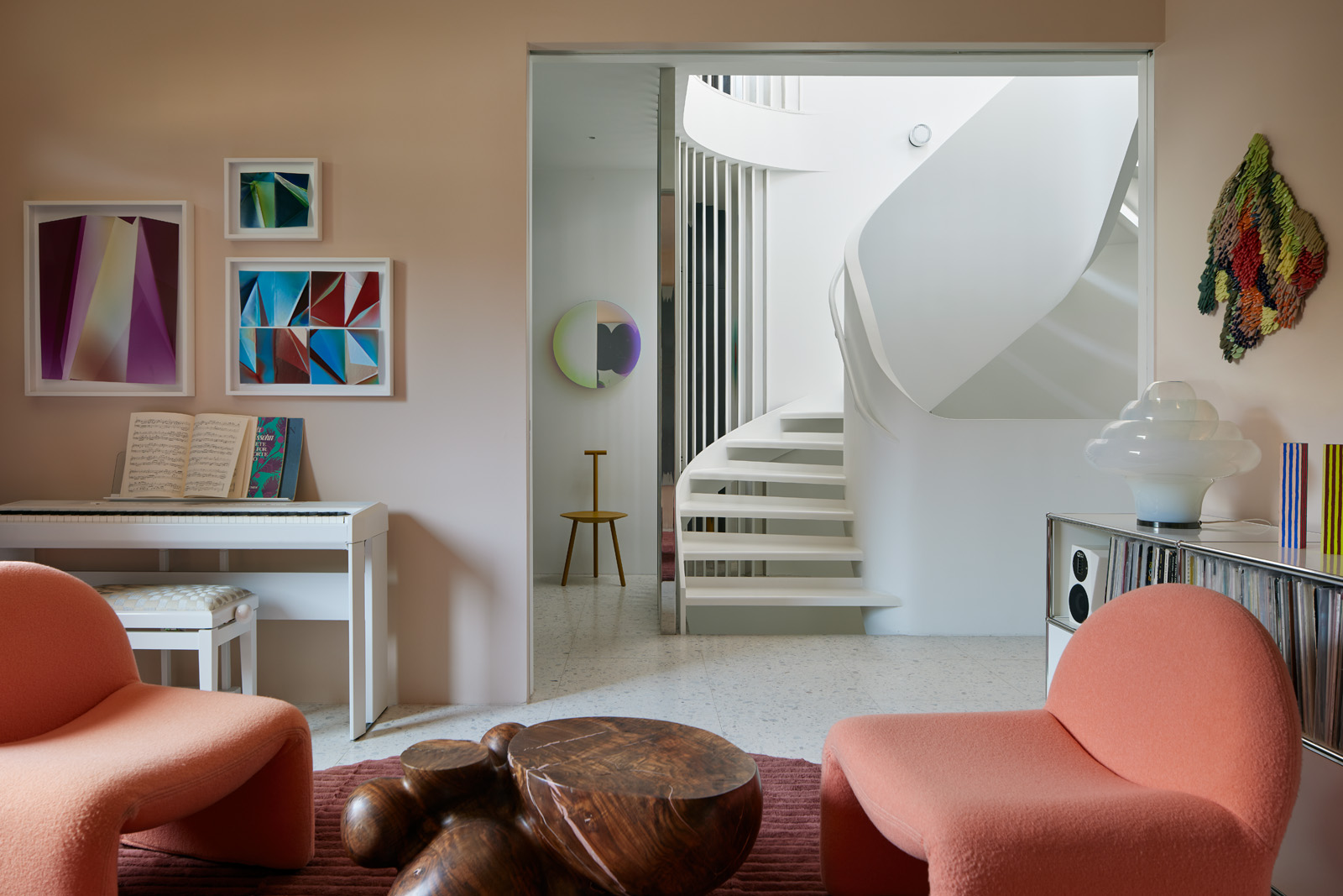 The room features two pink chairs, a sculptural wooden table, a piano with sheet music, colorful abstract art, a white spiral staircase, and modern decor.