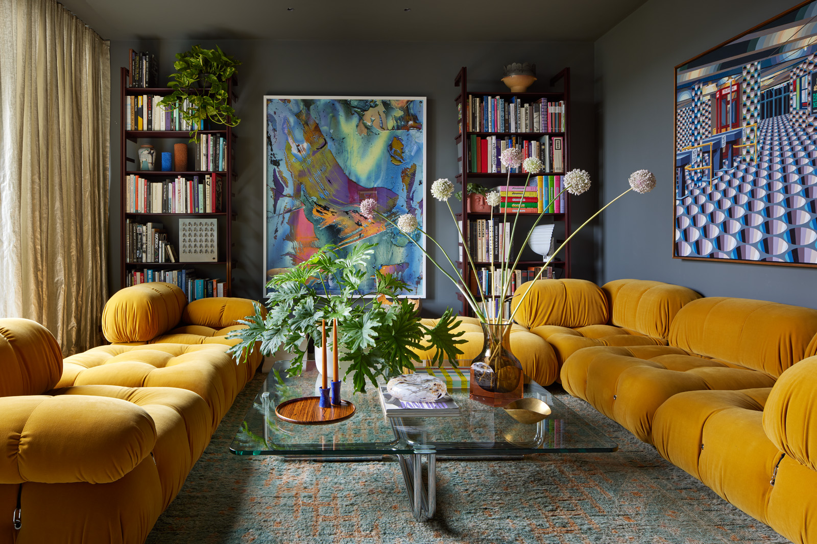 A cozy living room features mustard tufted sofas, vibrant abstract artwork, bookshelves filled with books, large plants, and a glass coffee table with decor items.