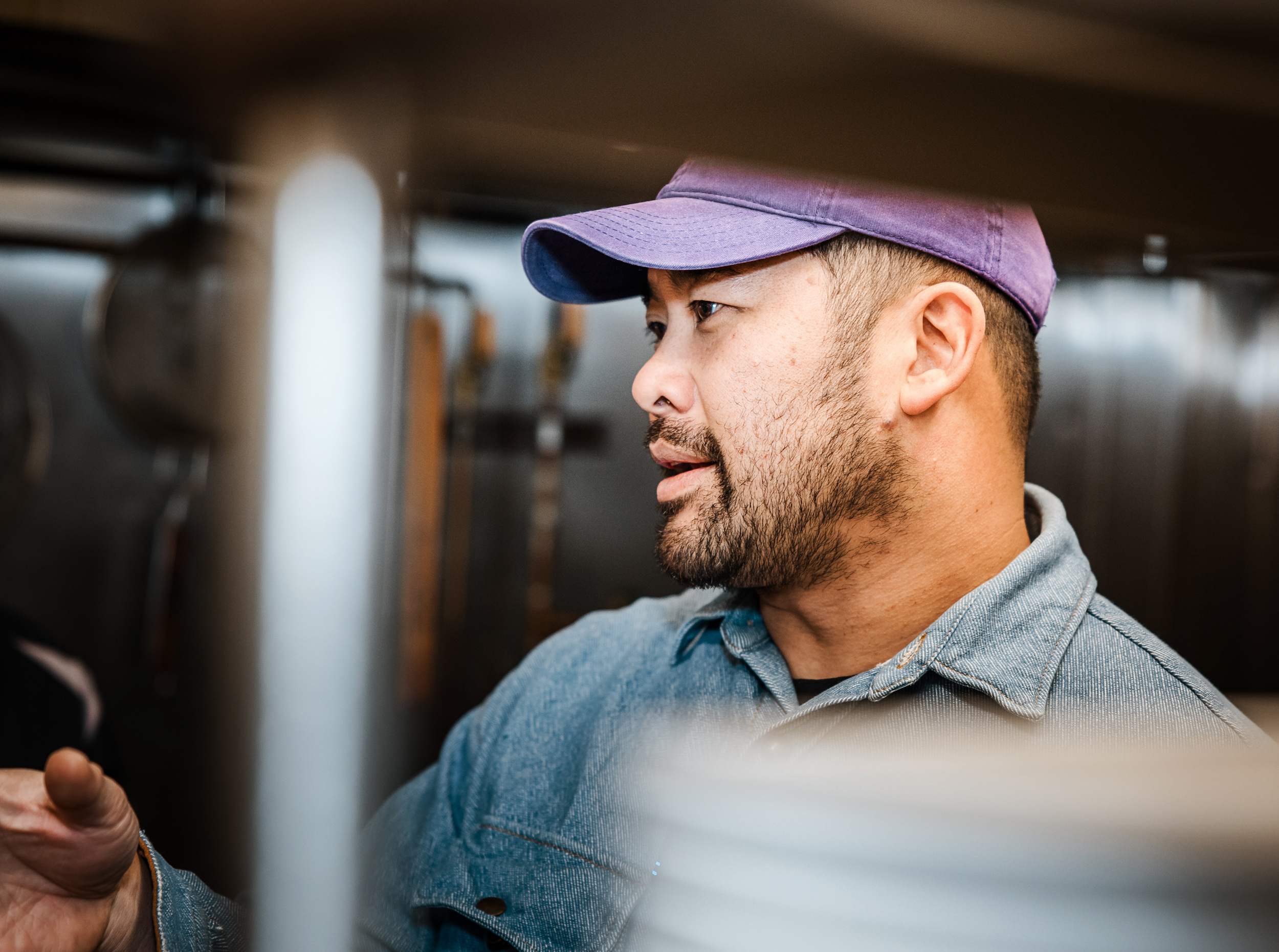 A man with a beard wearing a purple cap and denim shirt is focused on something, with a blurred kitchen background visible behind him.